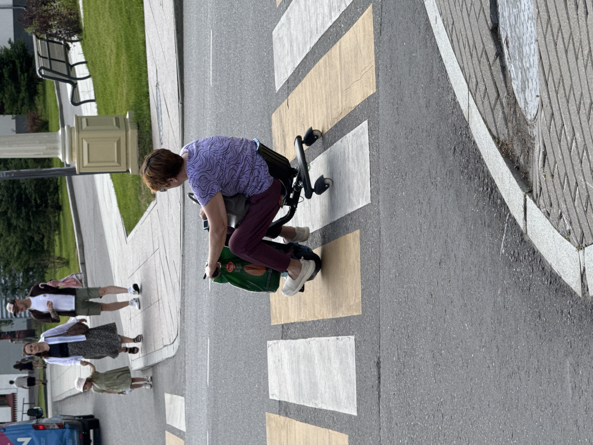 In Moscow, Russia, on a pedestrian crossing with yellow and white stripes, a woman with short red hair is riding a mobility scooter. She is wearing a purple t-shirt with a wave-like pattern and dark maroon pants. The scooter has black wheels and a black seat. She is carrying a large green shopping bag in her lap. In the background, a family is walking on the sidewalk. A woman in a black top and a patterned skirt, holding the hand of a young girl in a green dress and hat, walks past a man wearing a brown jacket, white t-shirt, and khaki shorts. The family appears to be walking away from the mobility scooter. The environment includes a paved road, a sidewalk, a green grassy area, and some trees.