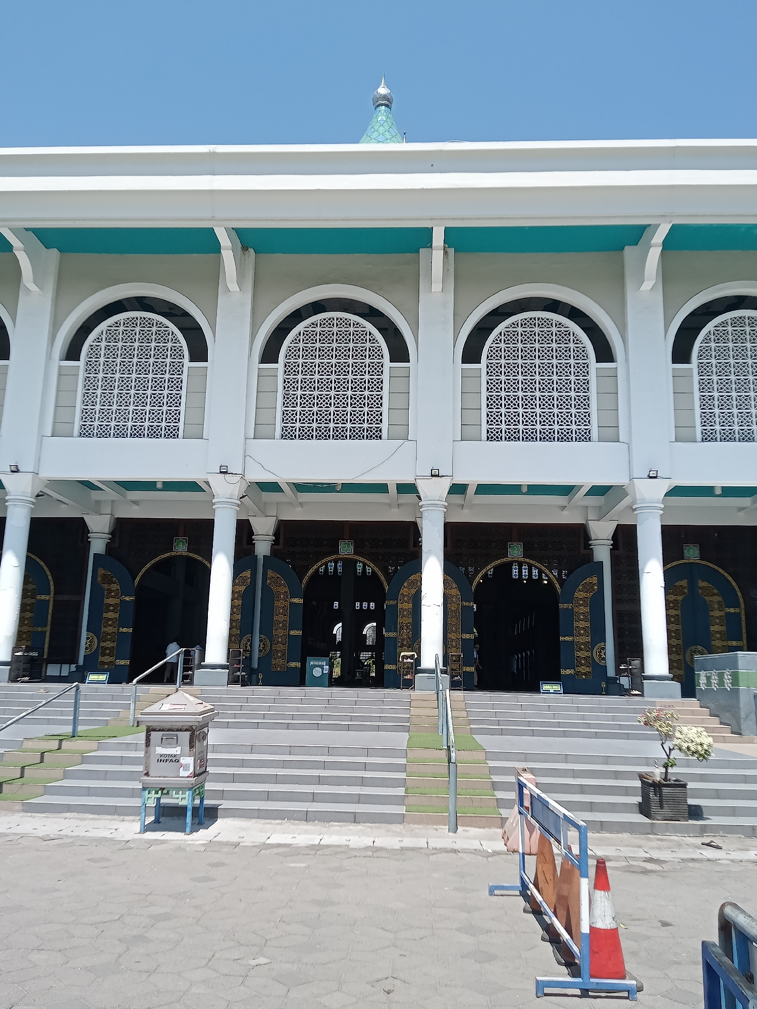 The image captures the facade of the Masjid Al-Akbar, a prominent mosque located in Surabaya, Indonesia, on a clear, sunny day. The mosque features a grand, multi-story structure with large, arched windows adorned with intricate white lattice work. A prominent turquoise band runs horizontally across the upper portion of the building, beneath a white cornice. Several tall, white cylindrical columns support the building's overhang, leading to the mosque's main entrance. Multiple dark-colored, arched doorways with ornate gold detailing are visible, suggesting the main prayer halls. In front of the building, a wide set of gray stairs ascends towards the entrance. To the left of the stairs, a metallic donation box labeled "KOTAK INFAQ" rests on a stand. Further down, a temporary barrier with traffic cones and a plastic bag suggests ongoing construction or maintenance. A small potted plant with white flowers sits to the right of the stairs, near a planter box. The sky is a vibrant, cloudless blue.