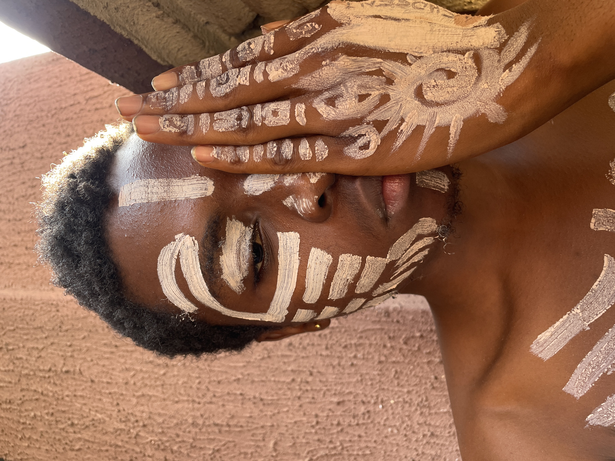The media file is a close-up portrait of a person with dark skin, adorned with white and beige body paint, presented with a 90-degree clockwise rotation. The subject's head is positioned towards the left side of the frame, with their body extending downwards and to the right.
The person's hands are placed over their face, partially covering the forehead and eyes, though one eye is visible and looks directly towards the viewer with a calm expression. Both the hands and face are decorated with intricate patterns of light-colored body paint. On the back of the more prominent hand, a circular sun-like symbol with radiating lines is painted. The fingers display patterns of short horizontal stripes and dots.
On the visible areas of the face, bold stripes are applied to the forehead and cheeks, with vertical lines near the nose and around the visible eye. Short, curly dark hair frames the person's head. A portion of their chest or shoulder is visible on the right, also bearing broad strokes of the paint.
The image is captured in bright, natural daylight, indicated by the clear illumination and subtle shadows. The background is an earthy-toned, textured surface, possibly a wall or ground, suggesting an outdoor or rustic setting. There is no visible text in the image. The scene conveys a sense of cultural or artistic expression, likely within the context of Onitsha, Nigeria.