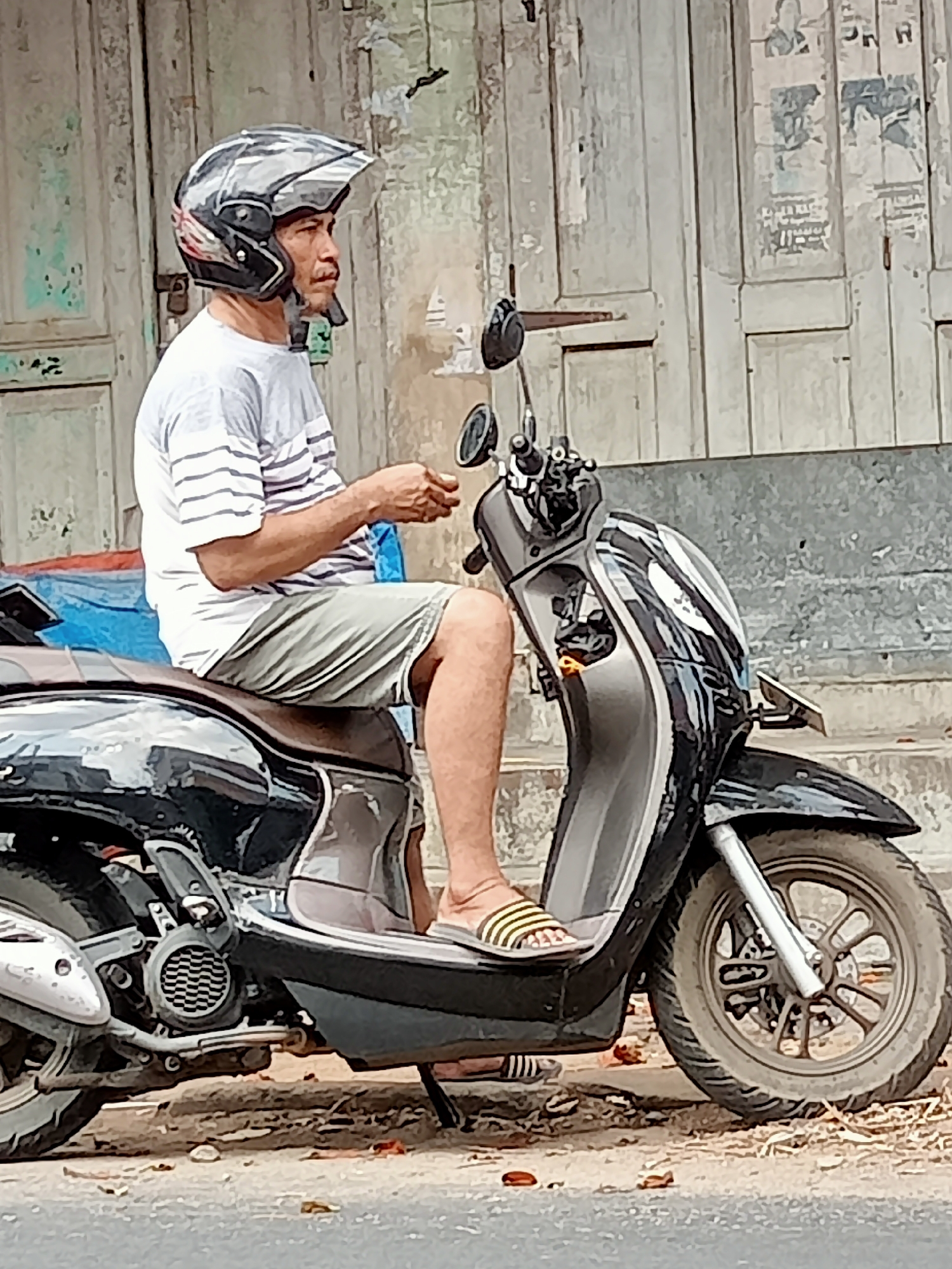A man wearing a helmet, a striped t-shirt, and shorts is sitting on a scooter parked on the side of the road in Tulungagung, Indonesia. The scooter is black with silver accents. Behind the man, a weathered wooden door with peeling paint is visible. The ground is covered with dirt, leaves, and small stones.