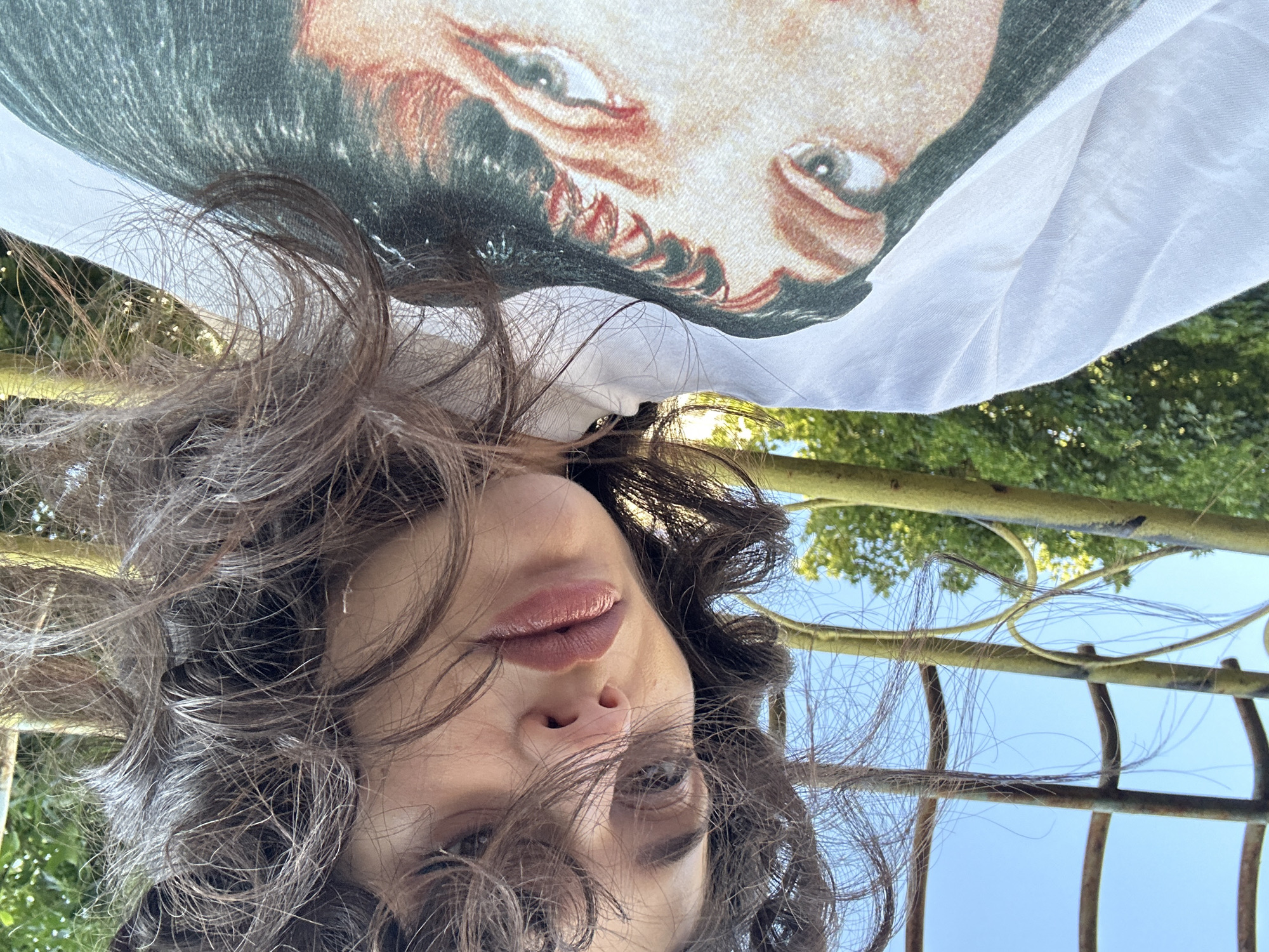 A close-up, low-angle shot captures a person's face and part of their white t-shirt, which features a graphic print of a person's eyes and face. The person's dark, curly hair is somewhat messy and obscures parts of their face. Their lips are slightly parted, and their eyes are visible, looking towards the camera. The background consists of a clear blue sky, green foliage, and a portion of a yellow metal structure, possibly a fence or garden trellis. The lighting suggests it is daytime. The location is identified as Sofia, Bulgaria.