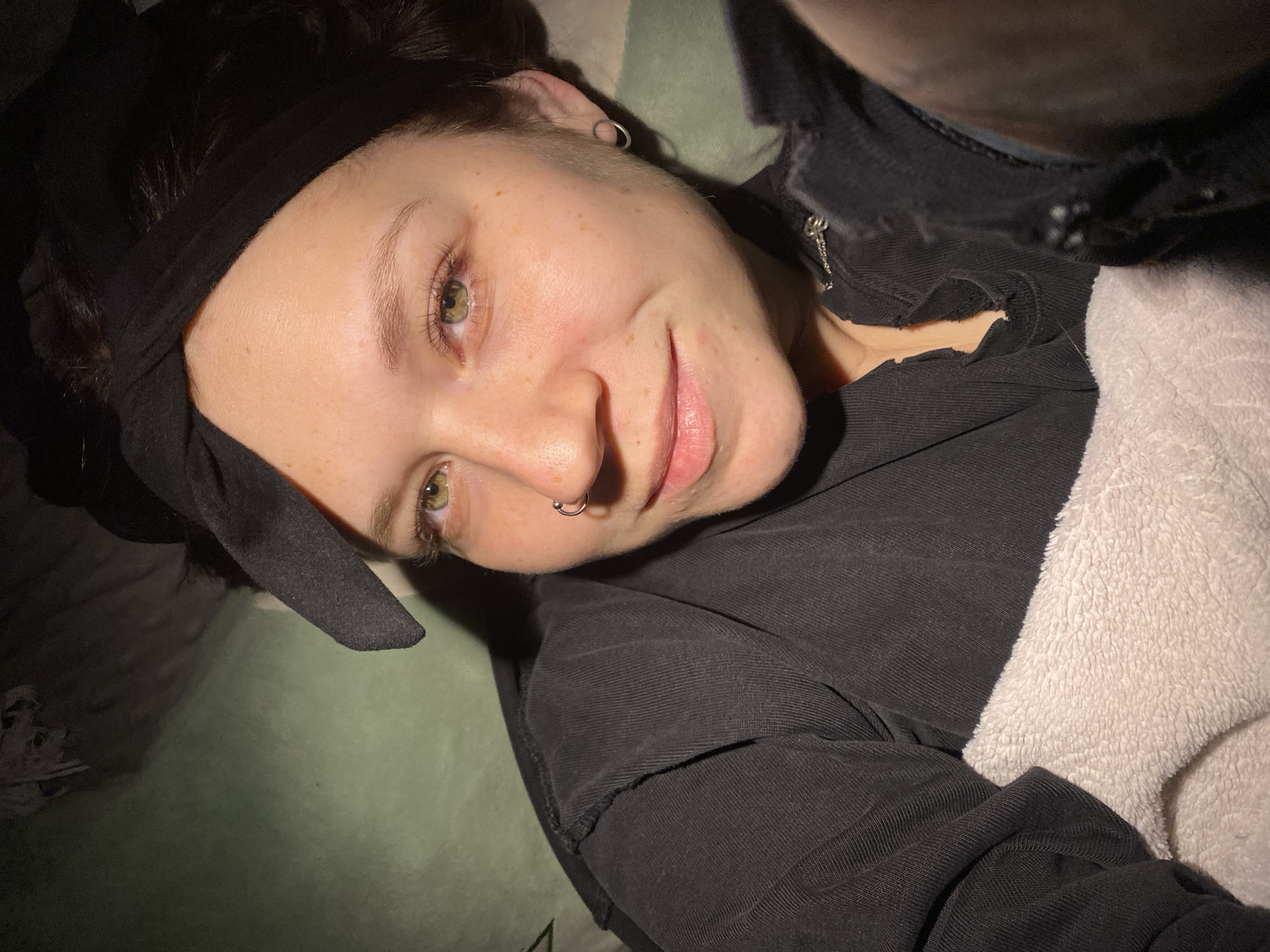 A close-up, low-angle, eye-level shot depicts a person lying down. The individual has fair skin, freckles, and light-colored eyes. They wear a black headband, a nose ring, and an earring. The person is dressed in a black top. A white, fluffy material is visible beside them. The background includes a green surface and glimpses of other indistinct objects. No evidence indicates the location is Chaikovsky, Russia.