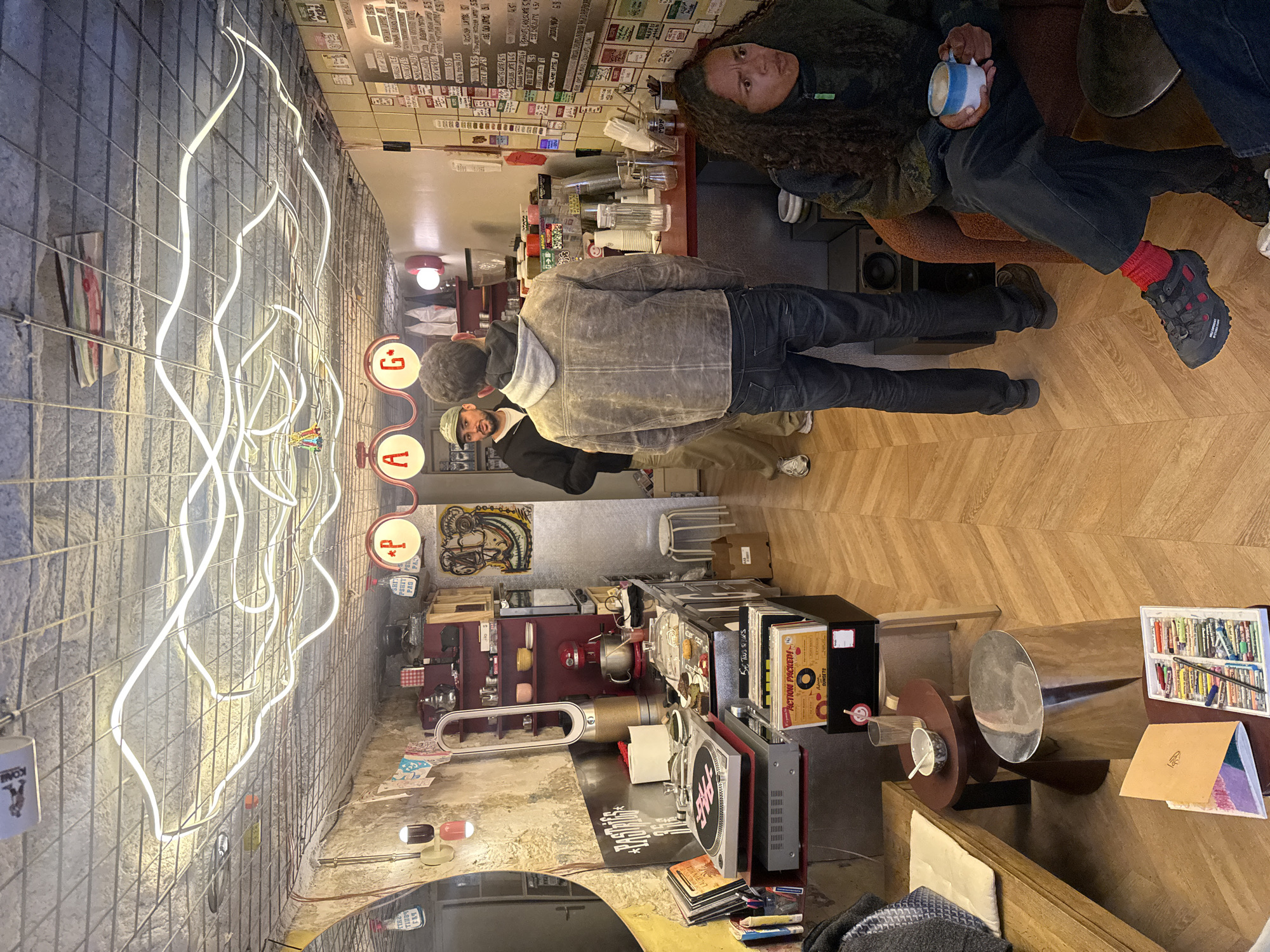 This is an indoor scene in what appears to be a cafe or small business in Paris, France. Three people are visible: one is sitting on a chair holding a mug, and two others are standing. The standing person is wearing a grey jacket and blue jeans. The location is crowded with various objects. On the right, there is a menu or list of items, and various other objects scattered around, including what appear to be vinyl records and art supplies. A distinctive neon light fixture is on the left wall.