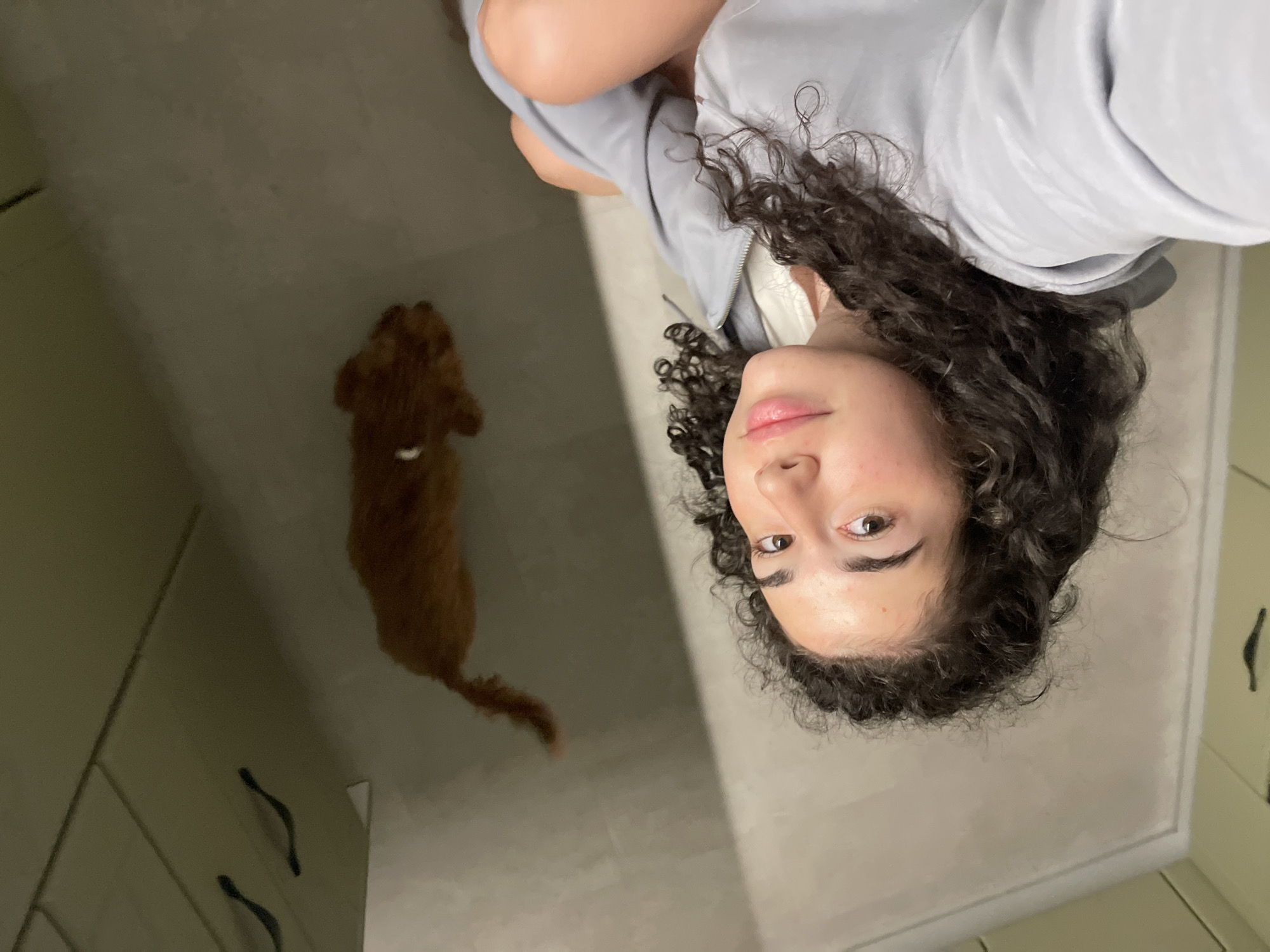 In a dimly lit indoor environment, a young woman with dark curly hair is taking a selfie. Her face, partially visible, shows eyes looking towards the camera and lips slightly parted. She is wearing a light gray hoodie. In the background, a small, reddish-brown dog, possibly a poodle or a similar breed, is lying down on the floor. The floor and walls appear to be made of a smooth, light-colored material, possibly tile or laminate. Above the dog, a section of cabinetry with dark handles is visible. The overall scene suggests a casual, domestic setting. The image does not provide any specific geographic indicators that would allow for its location to be determined as Sofia, Bulgaria.