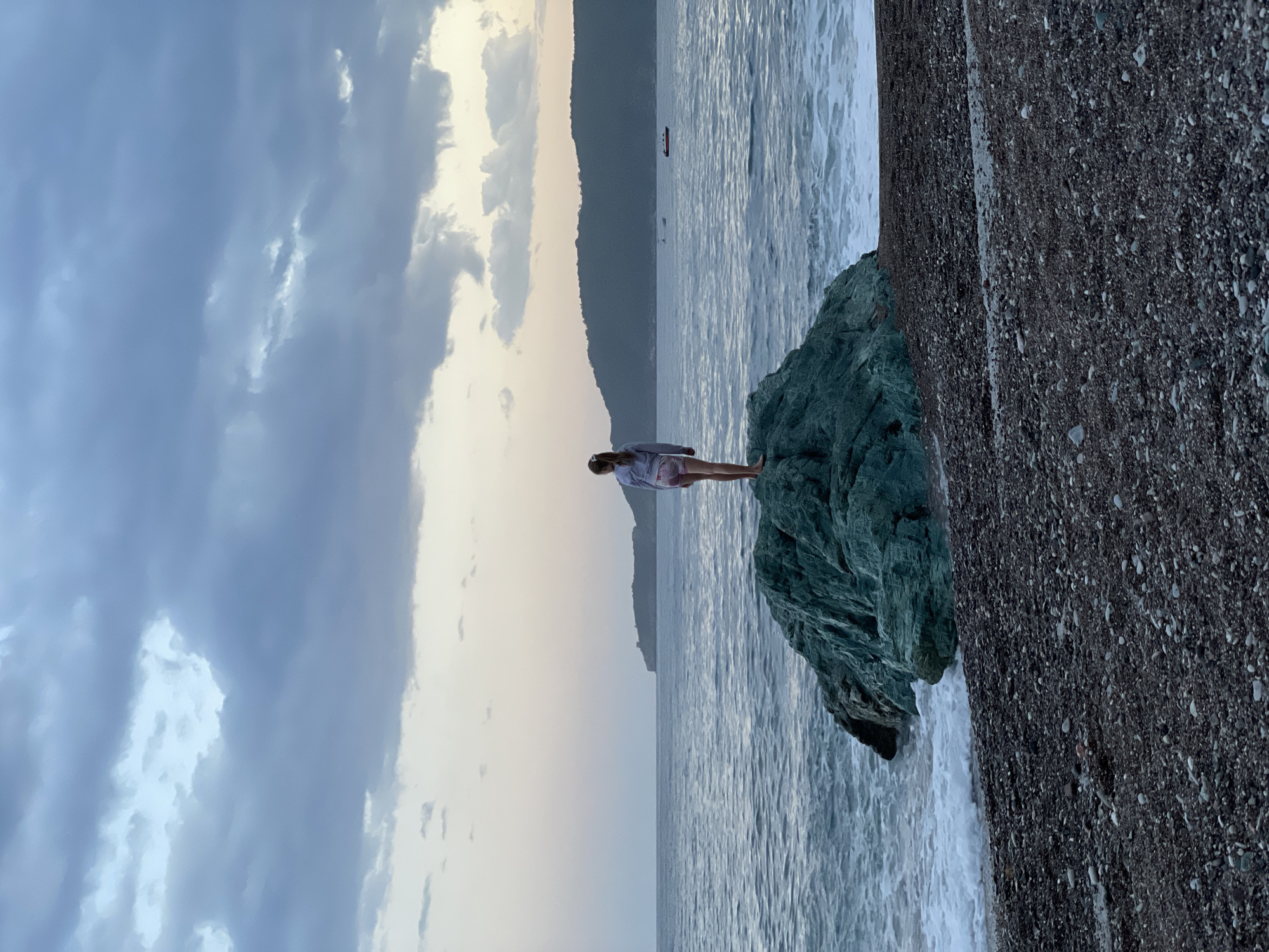The image shows a person standing on a rock formation at the edge of the sea, with a small boat in the distance and a mountain range across the water. The sky is overcast with clouds. The ground to the right of the rock formation is covered in pebbles.
Given the coastal environment, the overcast sky, the mountain range, the presence of rock formations, and the pebble ground, it is highly likely that the location is in Rafailovići, Montenegro.