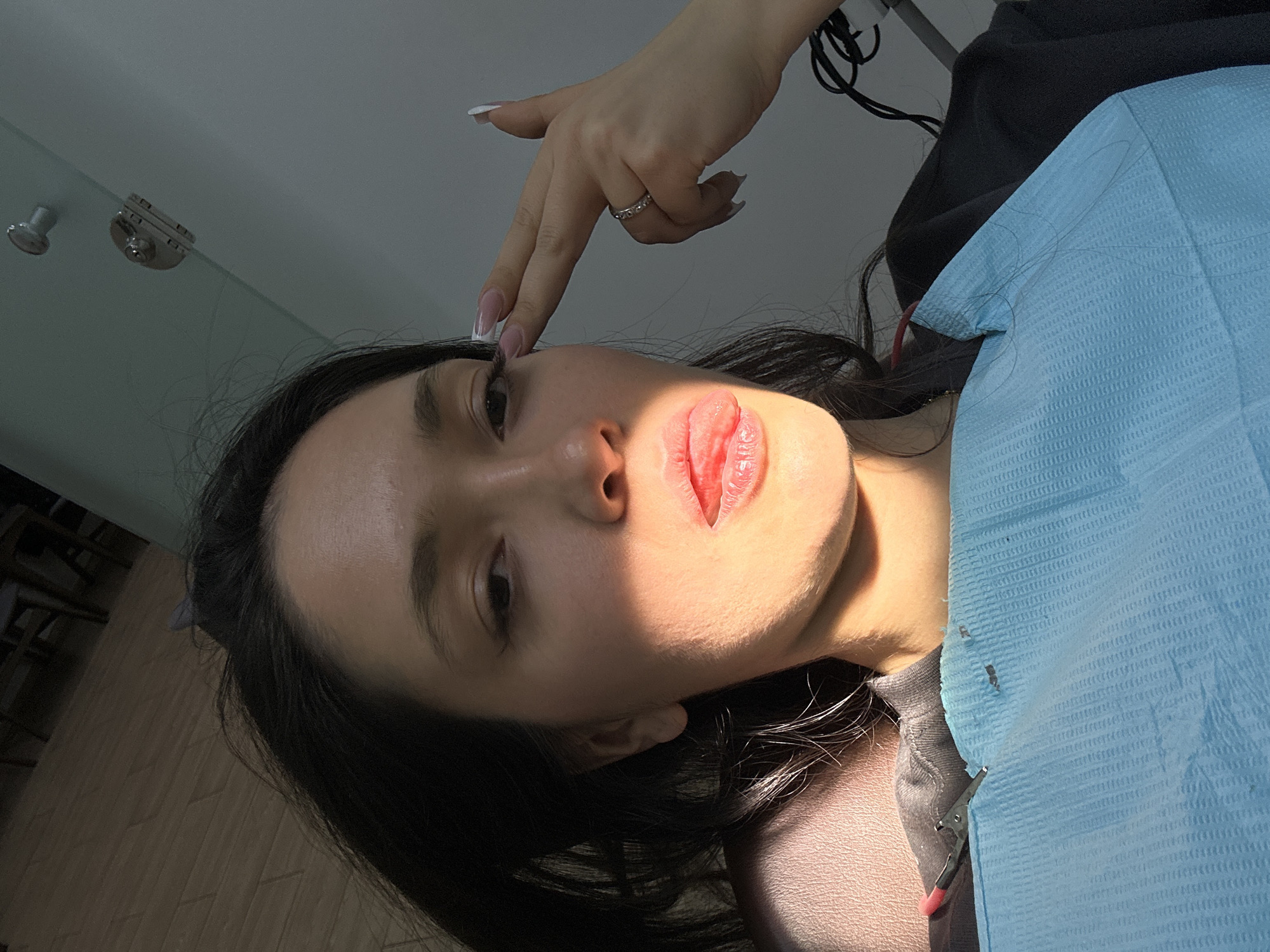 A woman lies on a dental chair, covered with a light blue bib, in a dental clinic in Yerevan, Armenia. Her dark hair is spread around her head. Another person's hand, adorned with a ring and manicured nails, touches her forehead. The scene is brightly lit. Dental equipment is visible in the background.
