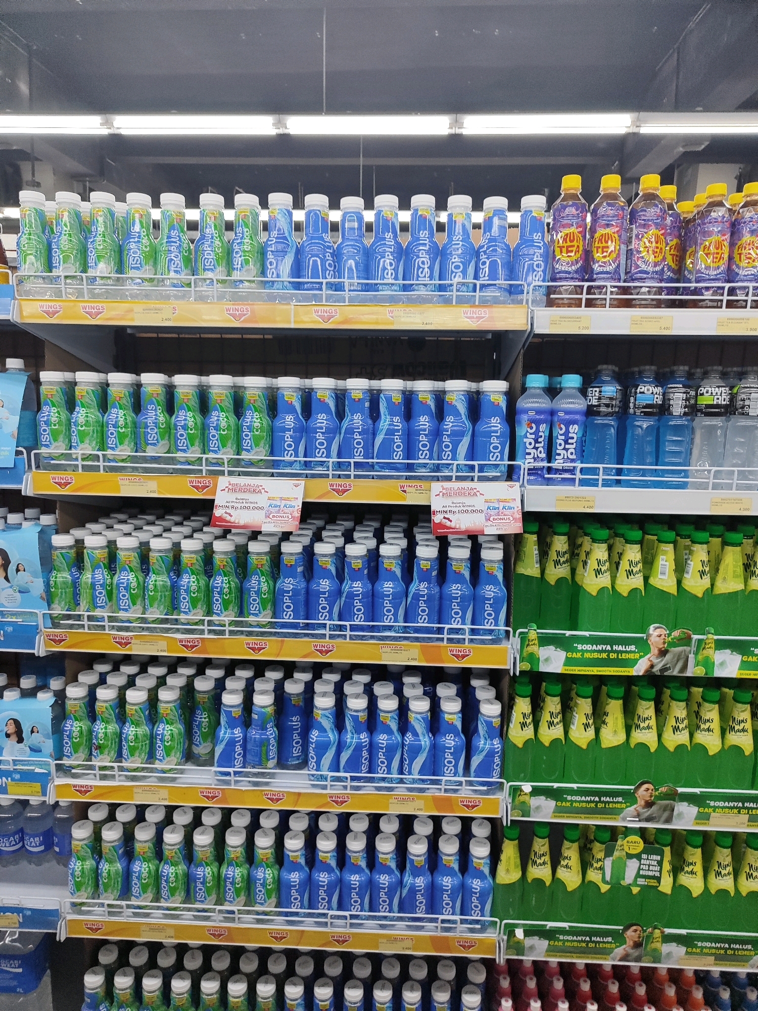 The image depicts a supermarket aisle stocked with beverages. The shelves are organized by brand and product type. On the top shelf, various flavors of Isoplus isotonic drinks are visible, with blue and green bottles predominating. To the right, there are bottles of Fruit Tea and what appear to be other flavored drinks with yellow caps. The middle shelves continue the display of Isoplus drinks, both coconut and blue variants. Also on the middle shelves, to the right, are bottles of Nips Madu (Honey Nips) in green packaging. The bottom shelves display more Isoplus drinks and then transition to rows of different colored bottles, potentially other types of soft drinks or juices. Price tags are visible on some shelves, indicating prices like 2,400 and 5,400. Promotional signage is also present, including one that reads "BELANJA MERDEKA" (Merdeka Shopping) with a bonus offer. The overall environment is brightly lit, typical of a retail setting. The specific location of Sukowiryo, Indonesia is not discernible from the visual information alone, but the product packaging suggests an Indonesian market. No people are clearly visible in the image.