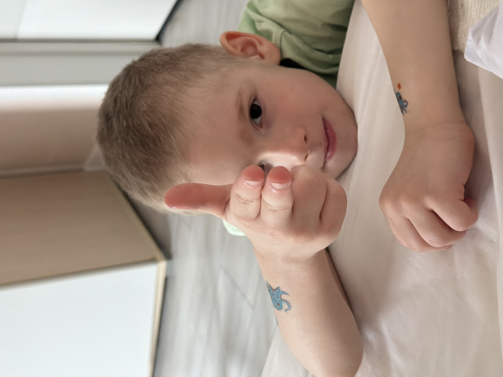A young child with short blonde hair is lying on a white surface. The child is wearing a green shirt. They are holding up their hand with their thumb extended. There is a blue tattoo on the child's forearm. The background includes a white wall and a glimpse of a gray floor. The location is Rossiyskiy, Russia.