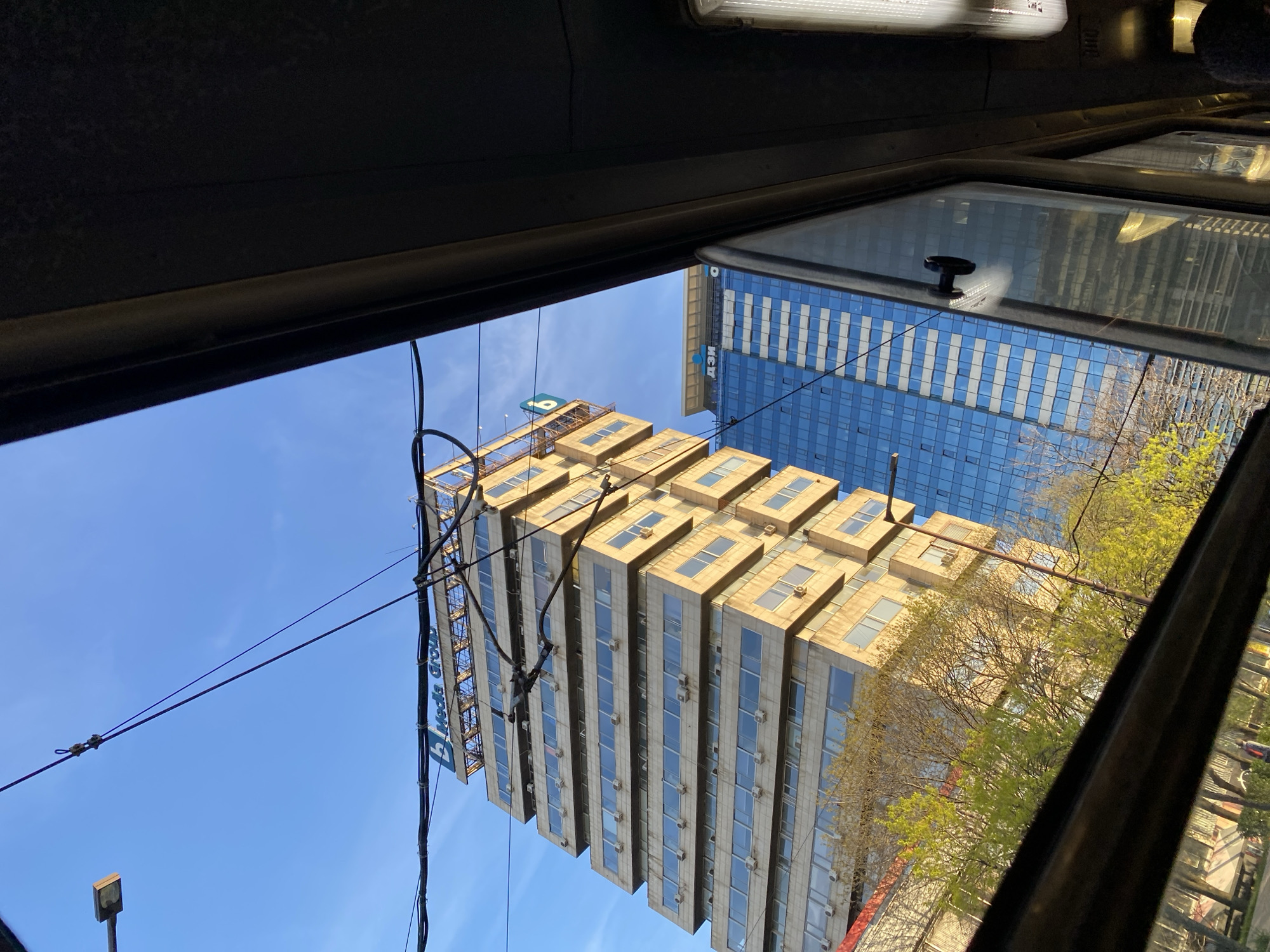 The image captures an urban cityscape in Sofia, Bulgaria, viewed from inside a vehicle, likely through a side window. The dark frame of the vehicle's interior is visible at the top and bottom of the composition. The setting is bright daytime under a clear, vibrant blue sky, suggesting sunny weather. The presence of trees with fresh, light green foliage indicates spring or early summer.
Two prominent high-rise buildings dominate the scene. On the left, a multi-storied building features a light beige or sandy-colored facade with a distinctive stepped or terraced architectural design on its upper floors. Numerous rectangular windows are visible across its surface, and many small white air conditioning units are mounted beneath them. At the very top of this building, a large sign or scaffolding structure is present, displaying the blue text "Hieds Group" along with a blue logo, though parts are obscured by utility wires.
On the right, a modern skyscraper with a striking blue glass facade rises. Its reflective surface mirrors parts of the sky and the surrounding environment. Faintly visible on this blue building's facade are the Cyrillic letters "ДЗИ", corresponding to "DZI", a well-known Bulgarian insurance company.
Several black electrical or utility wires crisscross the sky, running between structures and a street lamp visible in the bottom left. A notable detail is the clear reflection of the blue glass building and green trees in the vehicle's window, which also features a black handle or latch. No people are visible, and the image captures a static moment of observing the city from transit.