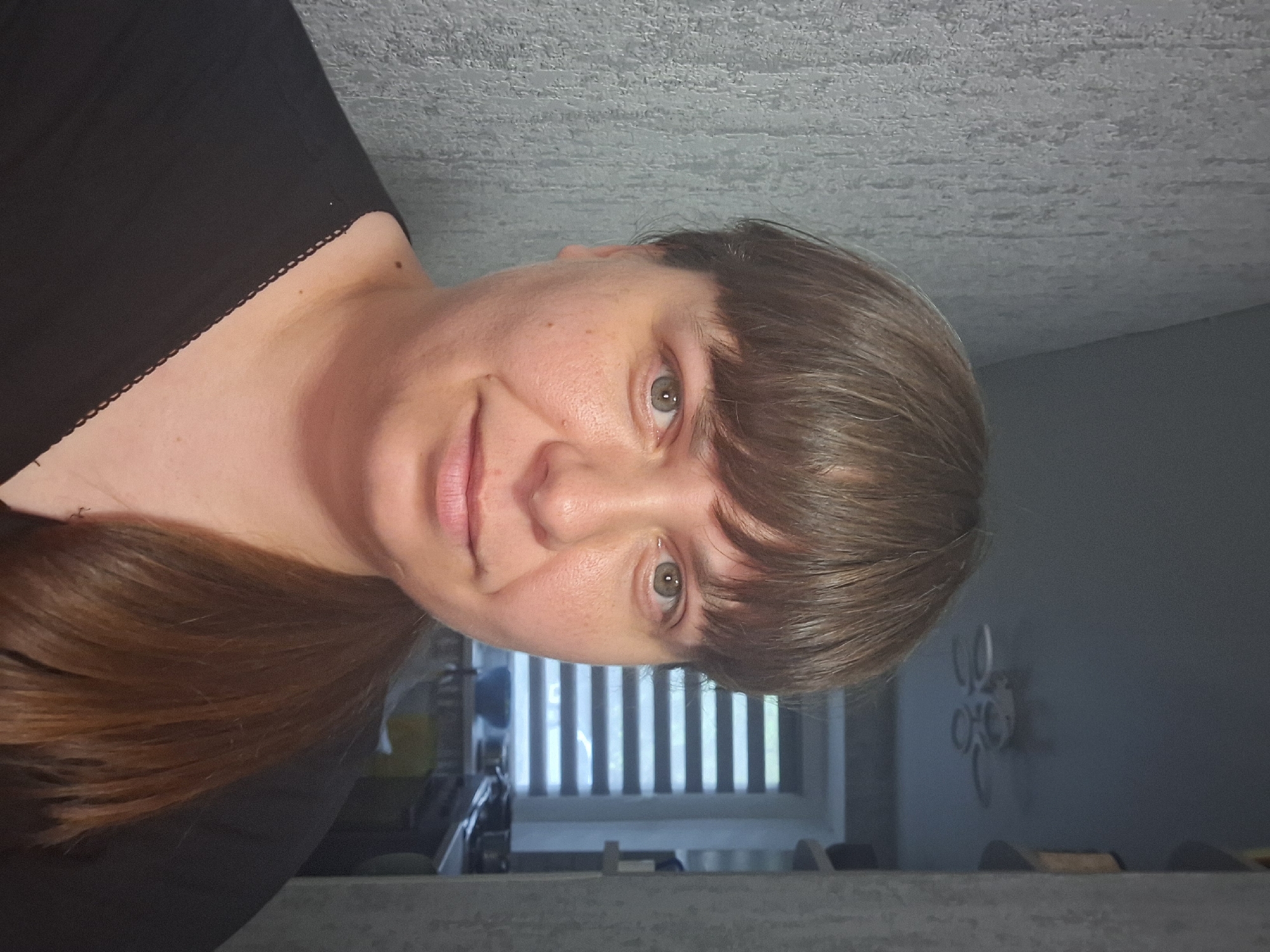 A medium shot, eye-level, selfie of a Caucasian woman with light skin, brown hair, and green eyes, taken indoors. She is wearing a black top with a decorative neckline. Her hair is pulled back into a ponytail. She has bangs and her hair is parted on the left. She is facing forward and slightly to her right, with a faint smile.
The background shows a wall with a textured pattern on the left, a ceiling with a fan, and a window with horizontal blinds on the right. The environment appears to be a domestic interior, possibly a kitchen or living area, based on the visible blinds and some out-of-focus objects in the background. The lighting is soft, suggesting an indoor setting. The image does not provide any specific clues about the exact location within Bila Tserkva, Ukraine, beyond it being an indoor residential space.