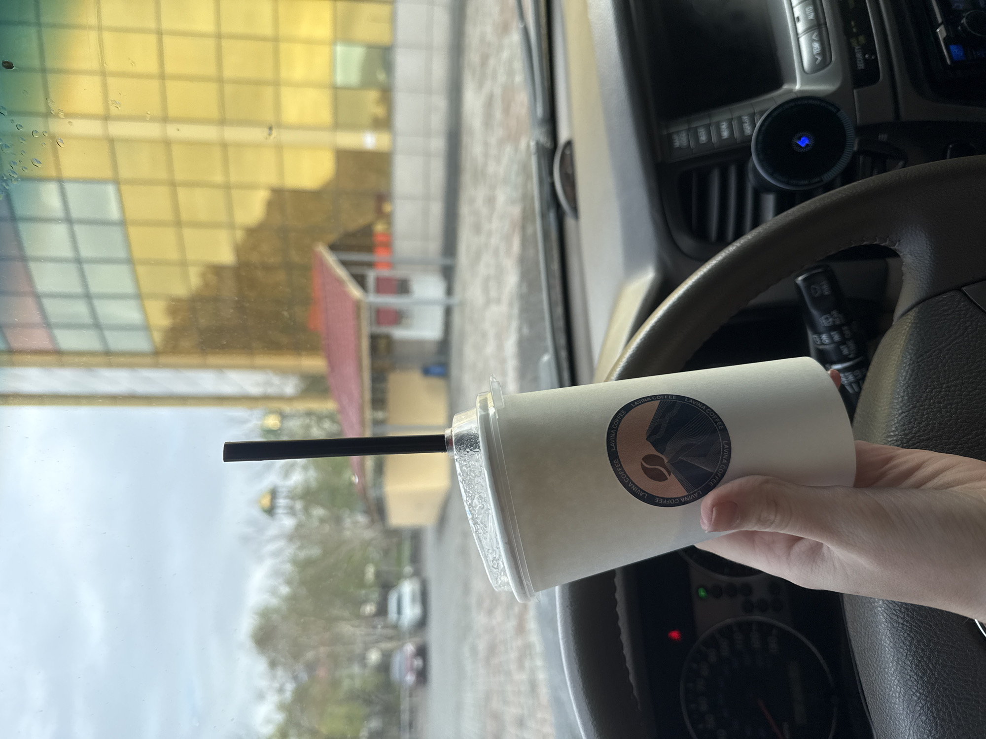 Here is a description of the image:
The image is taken from inside a car, looking out through the windshield. In the foreground, a hand holds a paper coffee cup with a black straw inserted. The cup has a white background and a logo that reads "LAVINA COFFEE." The car's interior, including the dashboard, steering wheel, and center console, is visible.
Outside the car window, the scene appears to be in Petropavlovsk-Kamchatsky, Russia. A building with a yellow and glass facade is visible, along with trees and a street with cars. The weather appears overcast.