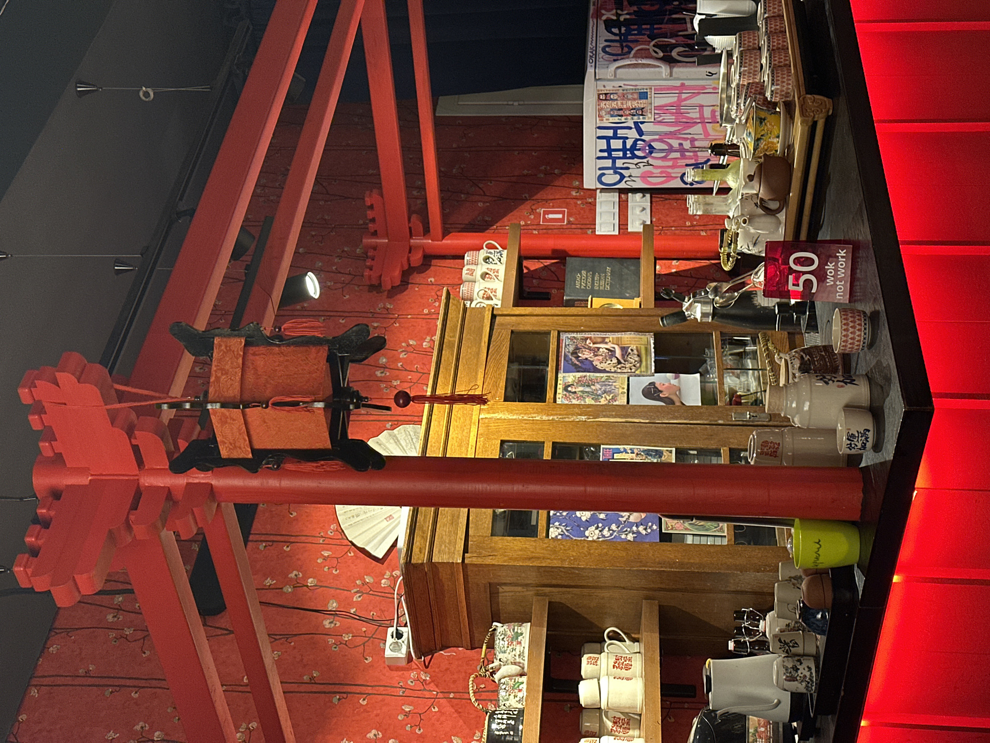 The image presents an interior scene, seemingly a shop or restaurant in Moscow, Russia. A torii-like red structure is prominently featured in the background, and its placement against the red floral wallpaper contributes to the overall aesthetic.
A wooden cabinet filled with various items, including books and ceramic mugs, stands adjacent to a countertop, which is laden with more mugs and decorative objects. A sign displaying "50 wok not work" is visible among the displayed items.
The scene is an amalgamation of cultural influences and objects, which creates an intriguing composition.
