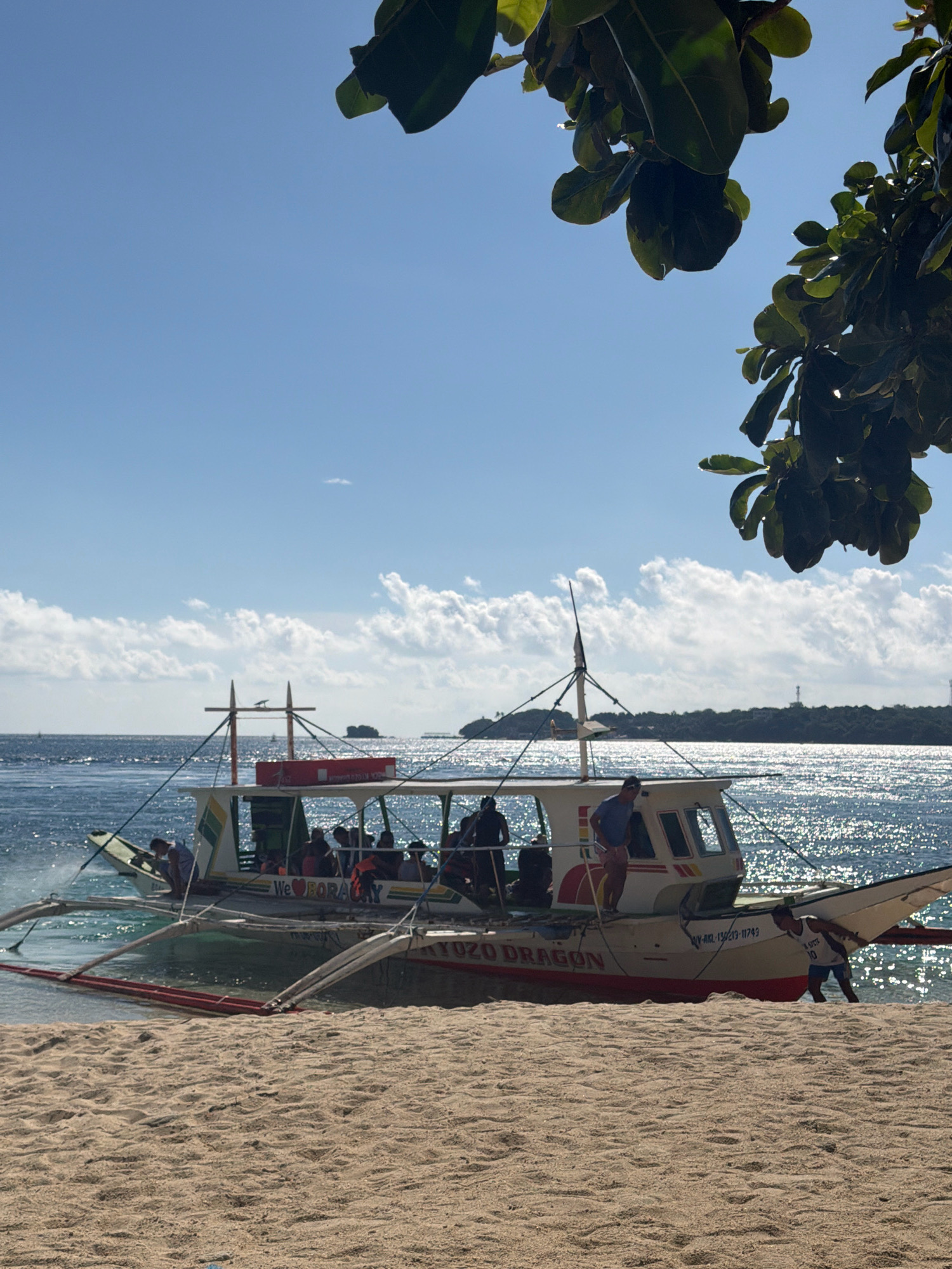 A bright, sunny day is depicted on a sandy beach in the city of Nabas, Philippines. A traditional Filipino outrigger boat, named "YAYO DRAGON," is beached near the shore, its outriggers extended. Several people are on board, some appearing to be tourists wearing life vests, while others are crew members. One crew member is visible on the sand, pushing the boat. The water is shimmering with sunlight, and a distant coastline with trees and structures can be seen across the sea. Lush green leaves hang from the top of the frame, partially obscuring the clear blue sky with scattered clouds.