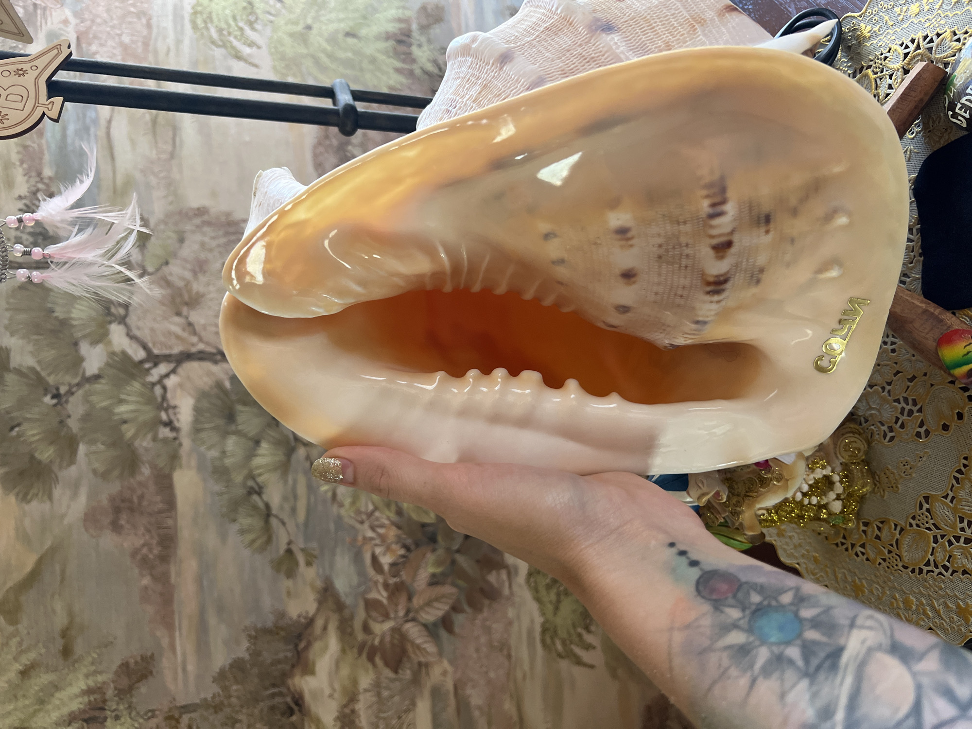 A person's hand, adorned with glittery nail polish, holds a large, iridescent conch shell. The shell is predominantly cream-colored with golden-brown markings and has a smooth, glossy interior. A faint gold inscription reading "SOCHI" is visible on the shell's exterior. Behind the shell, a decorative tapestry features a muted landscape with trees and possibly a waterfall, rendered in shades of green, brown, and cream. To the left of the shell, a black metal rack holds wooden ornaments and pink feathers with beads. The background also shows a glimpse of a table covered with a gold lace tablecloth, a small wooden piece with black lettering, and a colorful object. A tattoo is visible on the person's forearm. The lighting suggests an indoor setting, possibly a home or shop, with soft illumination.