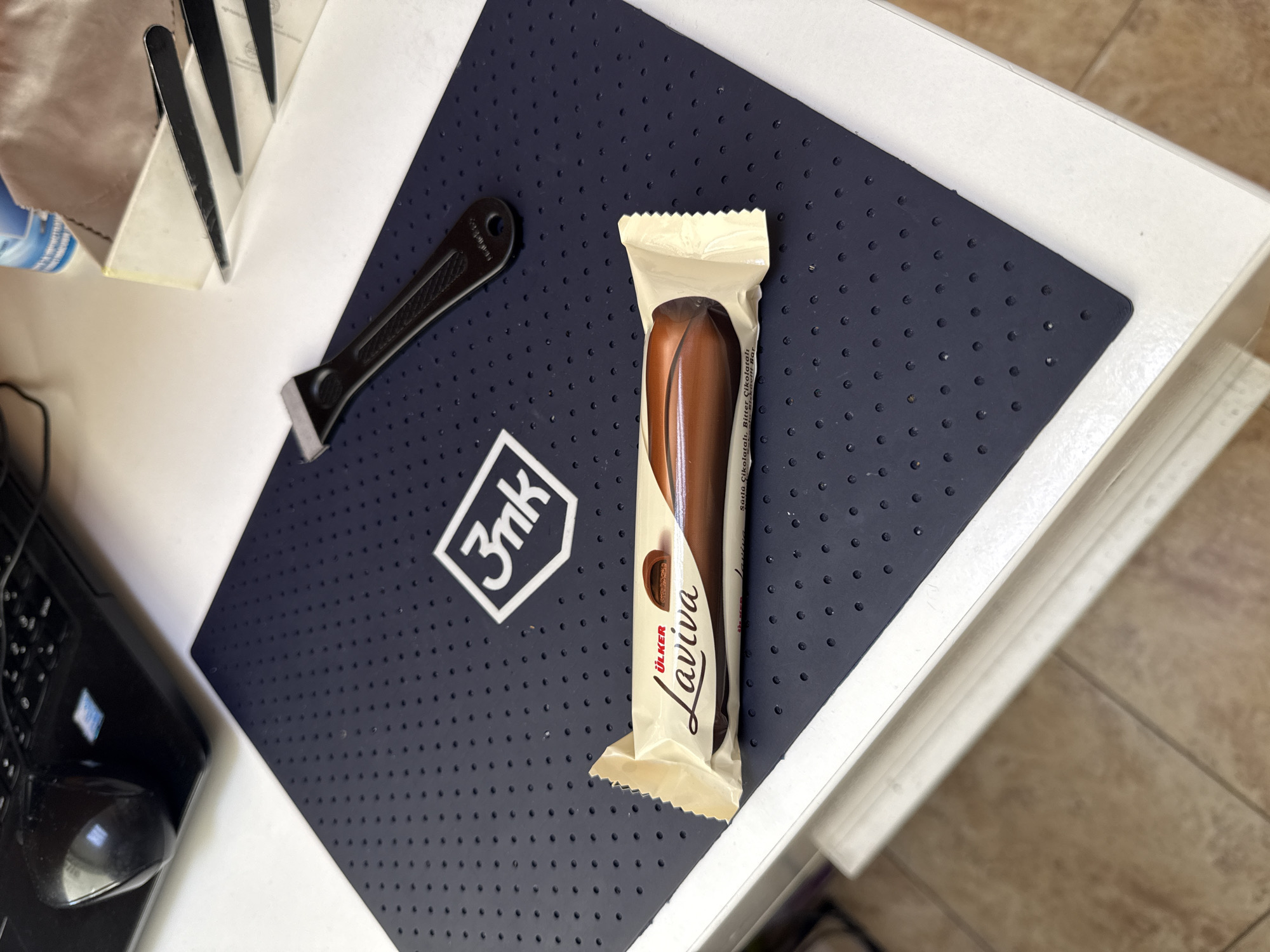 The image shows a still life scene on a white table with a dark blue rubber mat, which has a pattern of small circles. On the mat, there is a packaged Ülker Laviva chocolate bar with a white wrapper, a small black putty knife, and a white logo with "3nk" printed on it. Parts of other objects are visible in the frame, including part of a black computer mouse on the left, and a partially visible white object with some black strips at the top left. The scene is lit with natural light. The specific location in Burgas, Bulgaria is not discernible from the image.
