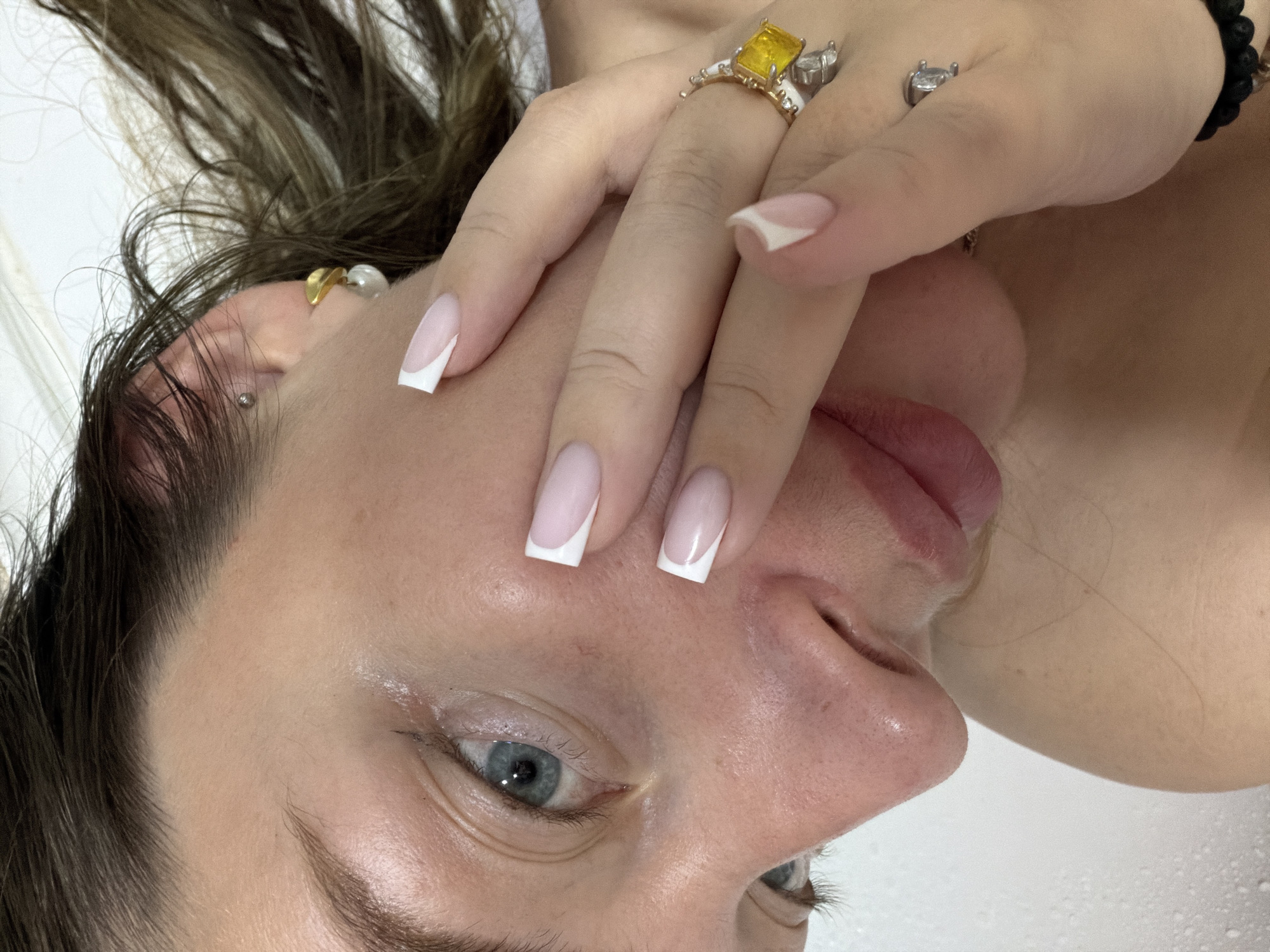 The image shows a close-up of a person's face, angled with their head slightly tilted. The person has fair skin, blue eyes, and brown hair with highlights. They are wearing multiple rings on their fingers, including one with a large yellow stone. Their nails are manicured with a French tip design. A portion of their shoulder is visible in the background. There is no visible information to indicate a specific location or to support the claim that the location is in the city of Chaikovsky, Russia.