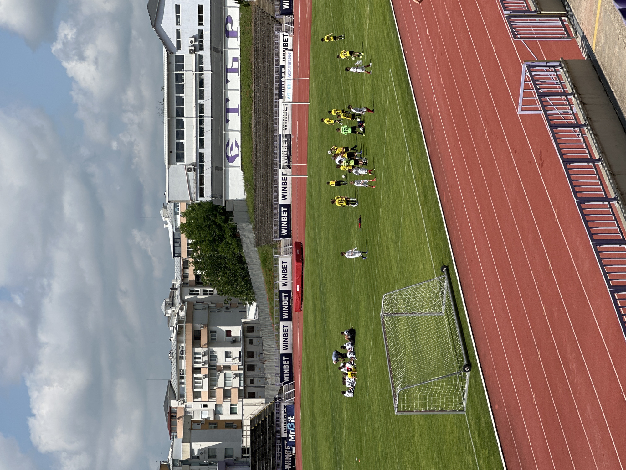 This image captures an aerial view of a football stadium in Veliko Tarnovo, Bulgaria, likely during a training session or a match on a bright, partly cloudy day. The scene shows a red running track with white lane markings, encircling a vibrant green football pitch.
In the mid-ground, a football goal with a white net is positioned on the right side of the pitch. Scattered across the field are numerous football players. A group of players in white and black striped jerseys, along with one in a white top and grey shorts, are gathered near this goal, some appearing to kneel or sit. Further up the field, a lone player in a white jersey stands. Closer to the center of the pitch, several players in yellow and green jerseys are engaged, some appearing to be in drills or small groups. A second, smaller goal is visible further up the field near these players.
Along the right sideline, behind the running track, are rows of empty stadium seating and a continuous line of advertising banners. These banners prominently display "WINBET" multiple times, along with "MrBit" and "NBET". The text "ЕКК АСКЛЕПИЙ" is also visible on a white banner alongside the stands. In the background, on the left side of the image, modern multi-story white and beige buildings rise beyond the stadium structures. Large purple letters, possibly reading "СТЛР" or similar, are visible on one of the white buildings. The sky above is a mix of bright patches and grey clouds. The overall scene depicts an active sports environment.
