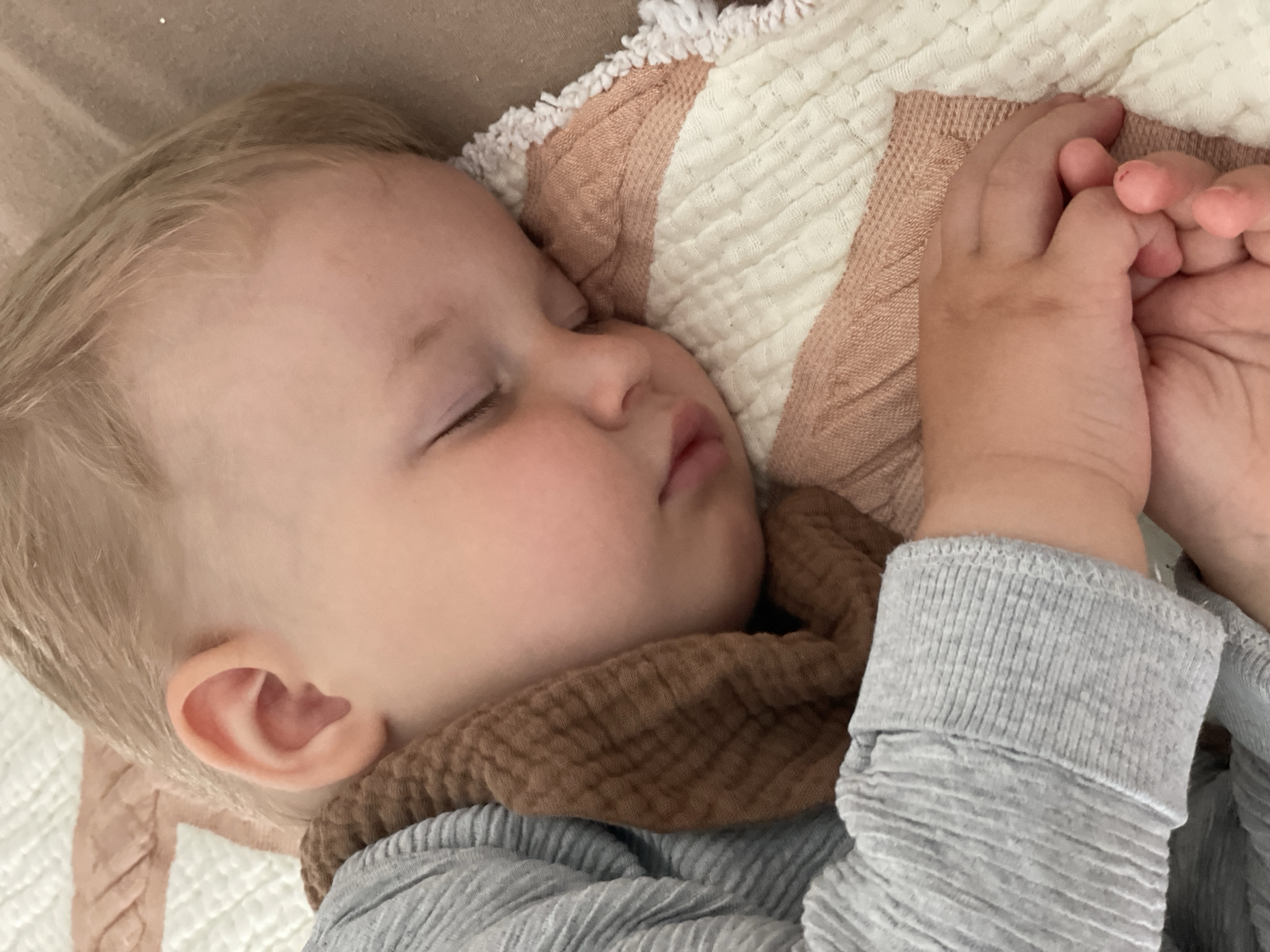 A close-up, eye-level shot shows a young child with blond hair sleeping on their side. The child is wearing a gray, ribbed long-sleeved shirt and a textured brown fabric around their neck, resembling a scarf or bib. Their head rests on a quilt with alternating horizontal stripes of white and light pink, fringed with white tassels. The child's right hand is visible, resting on the quilt, with fingers slightly curled. The background consists of a blurred brown surface, possibly a bed or sofa. The scene appears to be indoors, in a domestic setting, and is peaceful, capturing a moment of sleep. There is no identifiable location in the city of Nidda, Germany within the image.