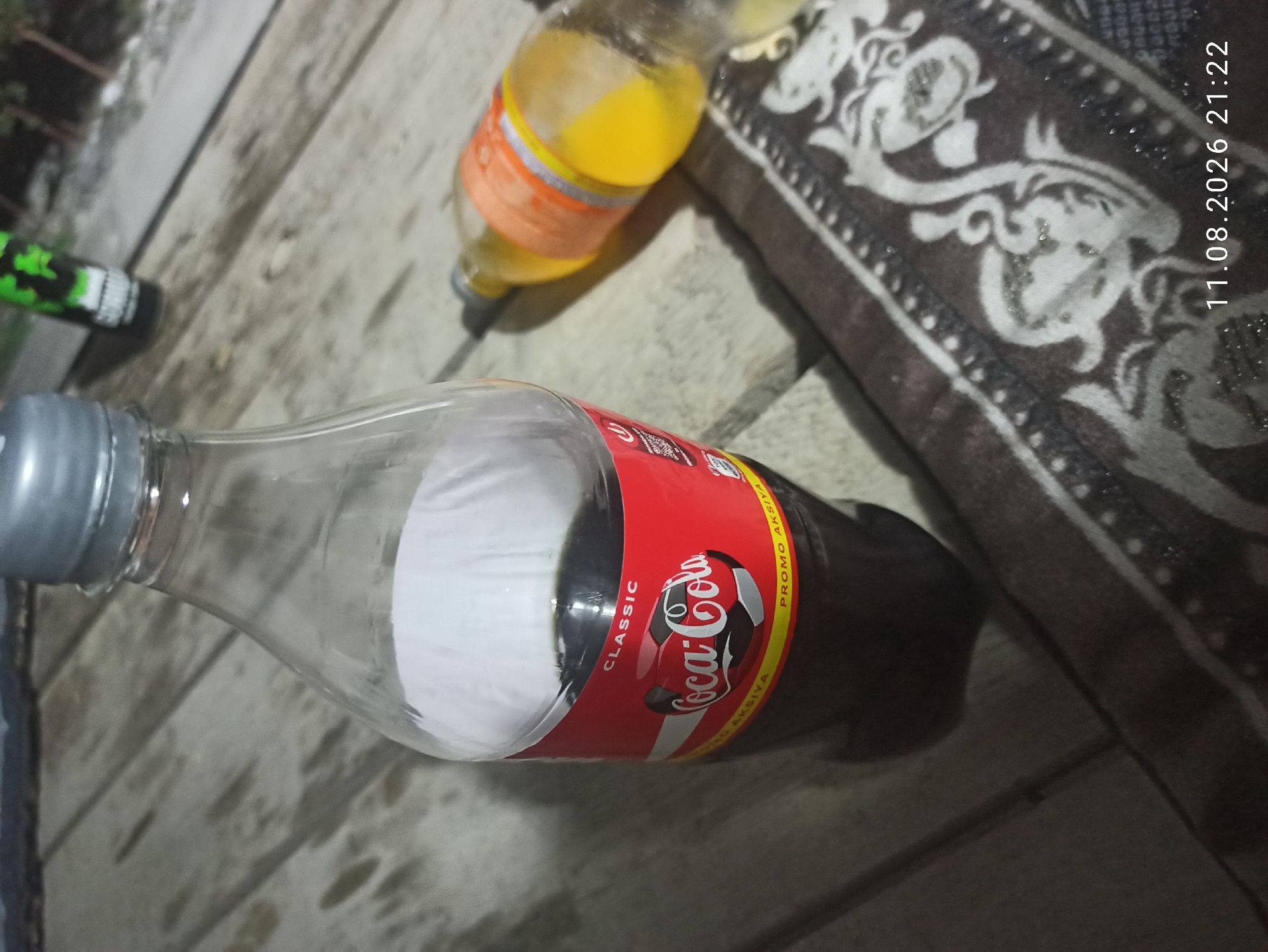 This is a close-up, slightly angled, low-light photograph taken at an outdoor table. In the foreground, a clear plastic bottle of Coca-Cola Classic is prominently displayed. The bottle is about two-thirds full of dark liquid, and its red label is partially visible, featuring the Coca-Cola logo with a soccer ball design. The cap of the bottle is silver. To the right and slightly behind the Coca-Cola bottle, another partially visible bottle, containing a yellow liquid, lies on its side. This second bottle appears to be a soda or juice. Both bottles are resting on a wooden surface that resembles planks of wood. A patterned textile, possibly a rug or blanket with a dark background and light-colored floral or paisley designs, is draped over the wooden surface in the lower right quadrant of the image. The background is out of focus, showing hints of greenery and possibly other objects. In the bottom right corner of the image, a timestamp is visible: "11.08.2026 21:22". The location is identified as Mulla, Uzbekistan, implying this scene is taking place within that specific city. The overall lighting suggests it is nighttime or dusk.