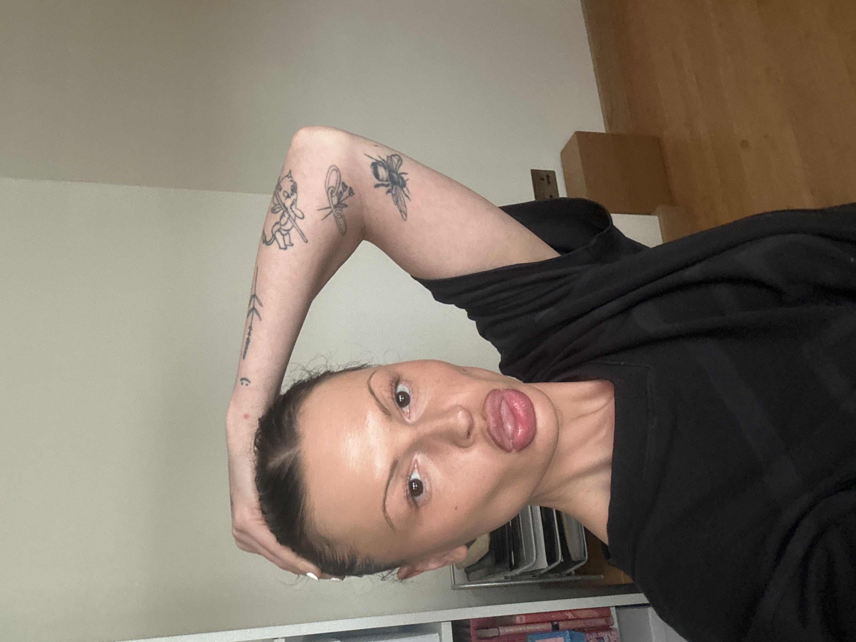 An indoor, head-and-torso level photograph depicts a female subject in Tunbridge Wells, United Kingdom. The subject has medium-brown hair pulled back, prominent cheekbones, and full lips with her tongue extended. Her right eye is clearly visible, brown in color. She is looking directly towards the camera. Her left hand, with white nail polish, is raised and positioned on her scalp near her hairline. Her right arm is bent and raised, displaying multiple black line-art tattoos, including a bee or insect, a floral motif, an arrow, and a small smiley face. A small red mark is visible on her forehead near her left hand. She is wearing a loose-fitting black short-sleeved top. The background on the left features a white bookshelf containing various books, some with reddish spines, and a multi-tiered metal document organizer. The background on the right consists of a plain light grey wall, with a light brown wooden block on the floor against the wall and a brown electrical outlet visible above it. The floor throughout the visible area is light brown wood.