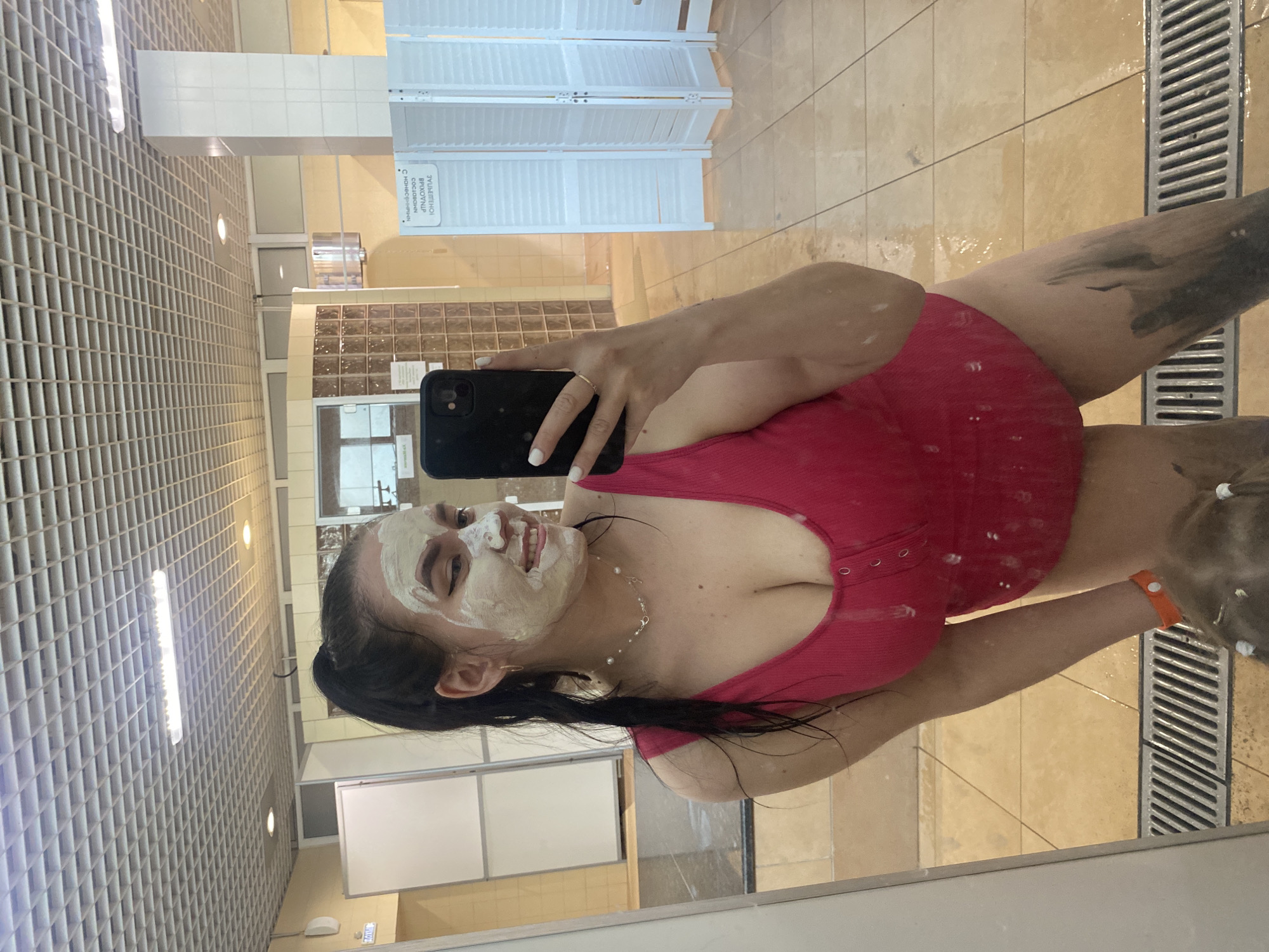 A young woman takes a selfie in a mirror inside a building in Votkinsk, Russia. She is wearing a red top and has a white facial mask applied to her face. Her arm and leg are covered in dark mud. She is holding a black smartphone in her left hand. The background includes a white tiled wall, a white folding screen, and a tiled floor. The building appears to be a spa or wellness center.