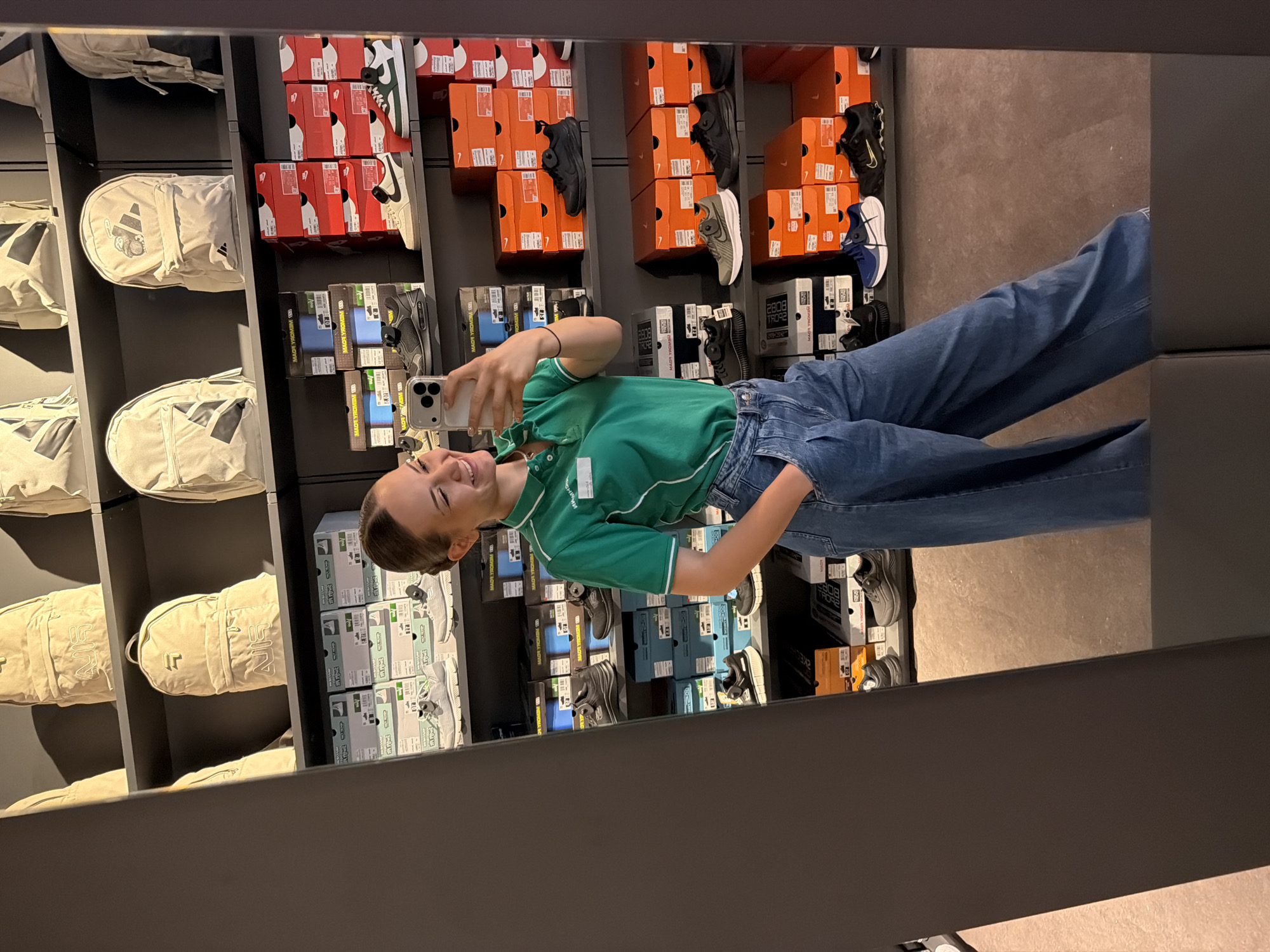 Here's a detailed description of the image:
**General Scene:**
The image shows a young woman taking a selfie in a store with a mirror. The store seems to be a sportswear or shoe store based on the merchandise on the shelves.
**People:**
* A young woman is visible in the mirror, holding a smartphone in her right hand and taking a picture of herself. She is smiling. She is wearing a green polo shirt and blue jeans. Her hair is styled back, possibly in a bun or ponytail.
**Objects:**
* Smartphone: The woman is holding a smartphone, presumably using it to take the selfie.
* Mirror: The woman is taking a selfie in a mirror that reflects her and the surrounding shelves.
* Shelves: Multiple shelves are visible, filled with various products.
* Shoes/Sneakers: Many shoe boxes from brands like Nike and possibly others are arranged on the shelves. Different shoe models are also displayed.
* Bags: Some shelves contain beige-colored Adidas bags and some other brands as well.
* Packaging Boxes: There are several boxes stacked on the shelves, likely containing shoes or accessories.
**Environment:**
* The environment appears to be a retail store. The lighting is relatively bright.
**Context:**
The photograph seems to capture a casual moment. The woman is possibly shopping or browsing in the store.
**Location in the City of Sofia, Bulgaria:**
It is not possible to determine the location in the City of Sofia, Bulgaria from the image.