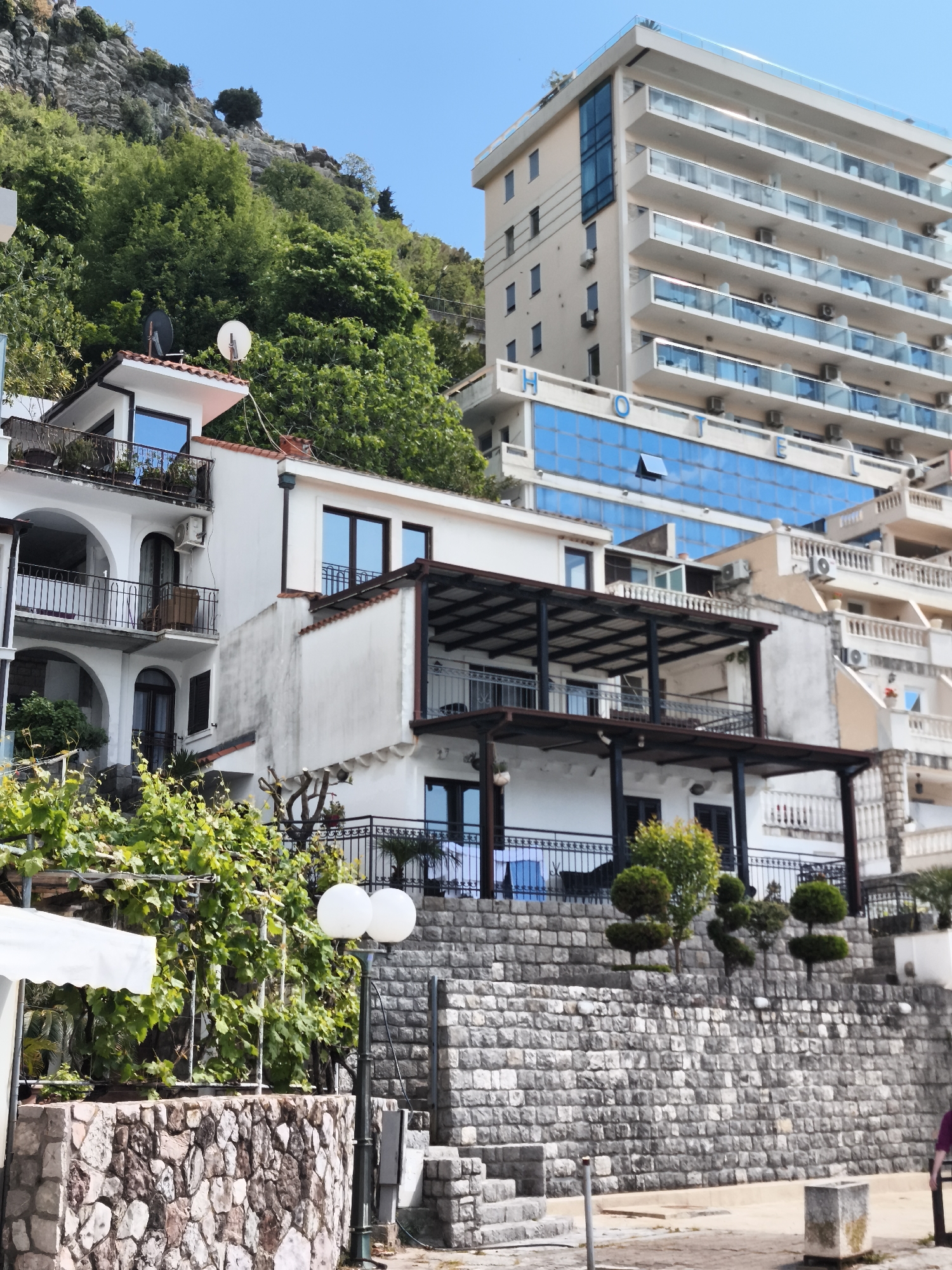 The image depicts a multi-tiered landscape in Rafailovići, Montenegro. On the left, a white building with arched doorways and wrought-iron balconies nestles against a lush, green hillside. Two satellite dishes are visible on its roof. Adjacent to it, a similar white building features a partially covered terrace with dark metal supports and railings. A retaining wall of gray stone bricks supports small, shaped trees. A taller, modern hotel building with multiple floors of balconies and blue glass sections stands behind these structures. The clear, blue sky completes the scene. A stone wall and foliage frame the foreground. A light post is also visible.