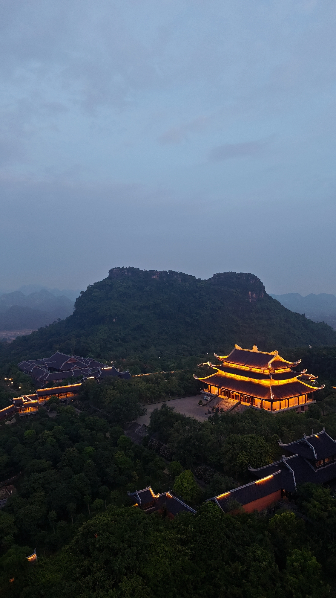 This is an aerial, high-angle, full shot of a temple complex at dusk in Tây Hoa Lư Ward, Vietnam. The sky is a soft, muted blue, with hints of pink in the distance. In the foreground, several traditional Vietnamese buildings with dark, curved roofs are illuminated by warm, orange-yellow lights. The largest building, centrally located, features intricate yellow trim and a multi-tiered roof. It sits amidst lush green trees, with pathways and other smaller buildings visible amongst the foliage. Behind the temple complex, a large, verdant mountain dominates the landscape. The mountain is covered in dense forest, with a rocky, flat summit. In the far distance, layers of misty, blue mountains recede into the horizon. The scene is peaceful and serene, capturing the grandeur of the temple architecture against the natural beauty of the Vietnamese landscape. There are no people visible in the image.