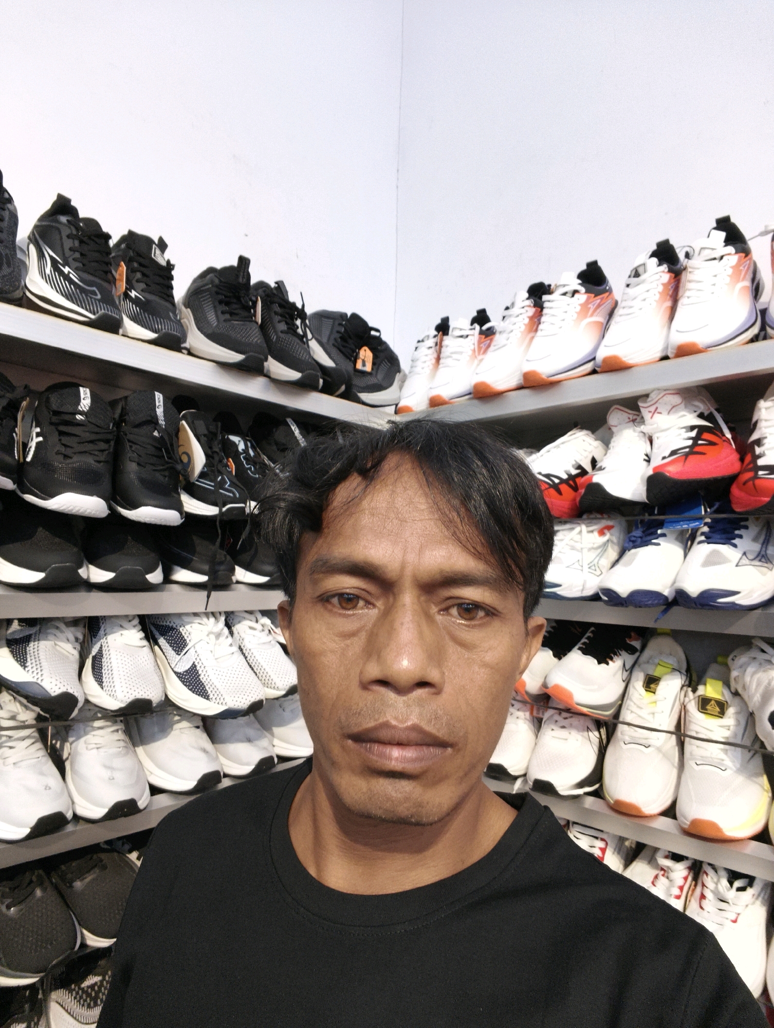 A man with dark hair and brown eyes is captured in a close-up, eye-level selfie, his face filling the lower-left portion of the frame. He wears a black t-shirt, and his expression is neutral. Behind him, a wall of shelves is densely packed with rows of athletic shoes, predominantly sneakers in various colors, including white, black, red, and blue. The shoes are neatly displayed in pairs on light-colored shelves, creating a visually repetitive pattern. The overall setting appears to be a retail environment, possibly a shoe store. Based on the provided location, this scene is situated in Ketapang, Indonesia.