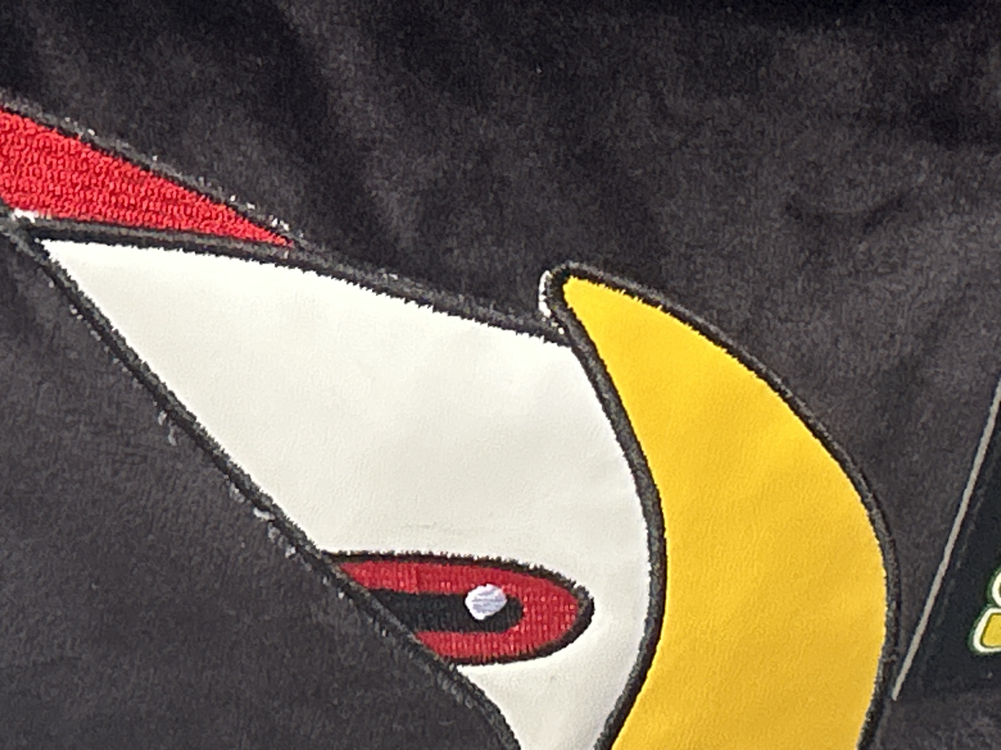 A close-up image shows a portion of an embroidered design on black fabric. The design appears to depict a stylized bird's head, with a red patch at the top, a white main area featuring a red eye with a white highlight, and a yellow section on the right. The fabric and embroidery have a textured appearance. The location, city, and other surrounding context are unknown.