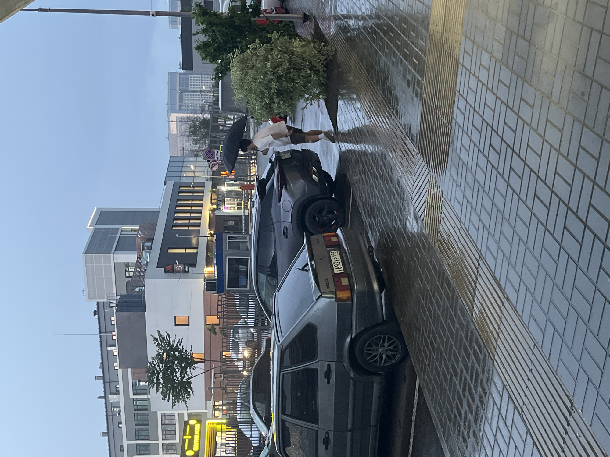 The image shows an outdoor parking area on a cloudy day, with rain evident on the wet pavement and surfaces. In the foreground, a gray Lada hatchback is parked. To the right of the Lada, a darker colored SUV is parked. A person wearing a white t-shirt and dark shorts is standing between the SUV and a large green bush, holding a black umbrella over their head. They are facing away from the camera, looking towards the SUV. In the background, several multi-story buildings of modern architecture are visible, with some windows illuminated. The pavement is made of dark rectangular bricks laid in a herringbone pattern, with a textured strip indicating a pedestrian crossing or tactile paving. The sky is overcast, suggesting poor weather conditions. This scene is consistent with an urban environment.