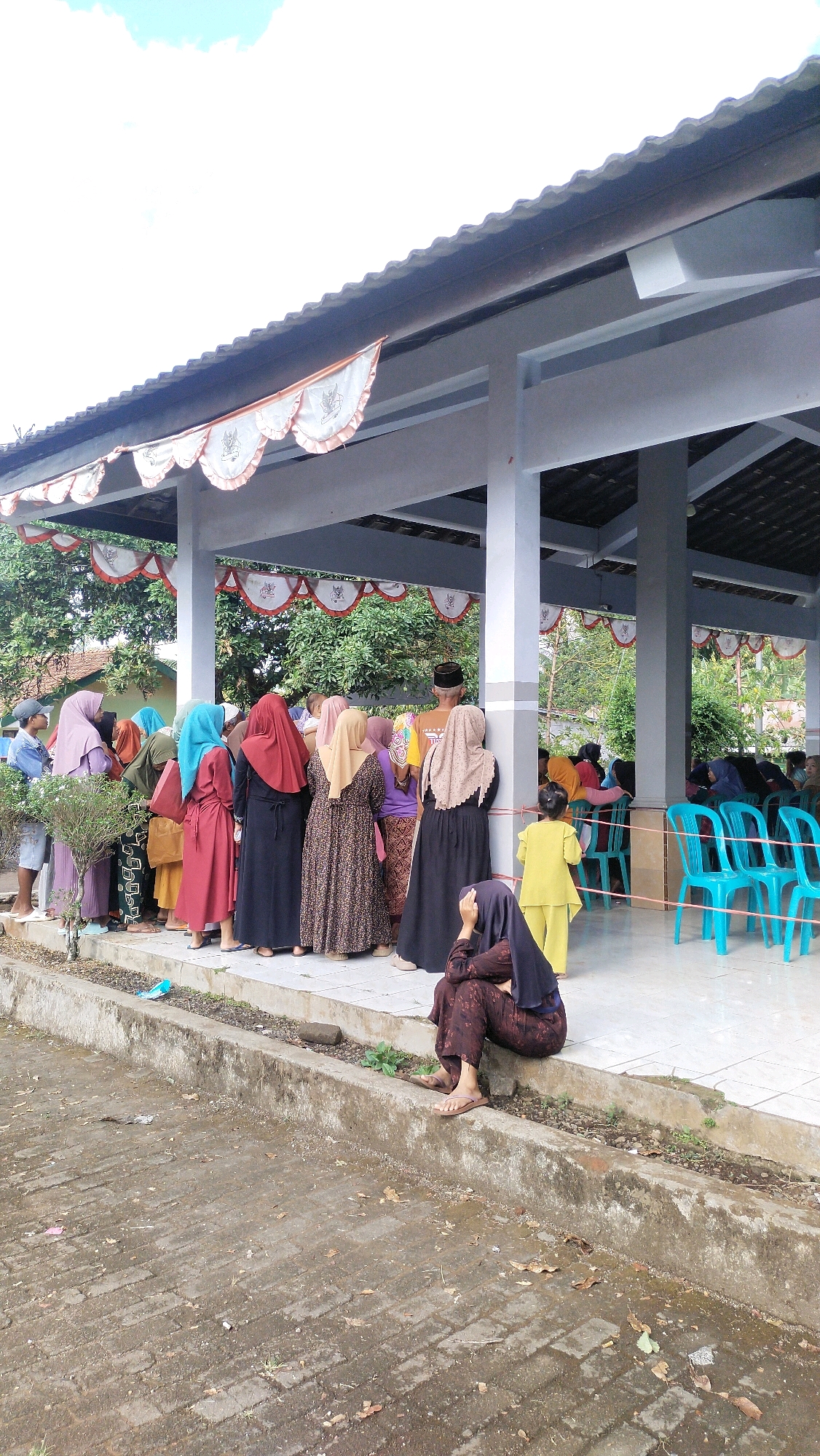 In Klungkung, Indonesia, a group of people are gathered under an open-air pavilion with a tiled roof, decorated with red and white pennants. The pavilion is supported by white pillars. A line of individuals, mostly women wearing hijabs and traditional Indonesian attire, stand facing away from the camera, appearing to be in a queue or attending an event. In the foreground, a woman in a dark hijab and patterned brown clothing sits on a concrete ledge, looking down and resting her hand near her face. She is wearing sandals. In the background, more people are visible inside the pavilion, some seated on blue plastic chairs. The ground is paved with rough concrete and scattered with dry leaves and debris. The environment suggests an outdoor community gathering or event.