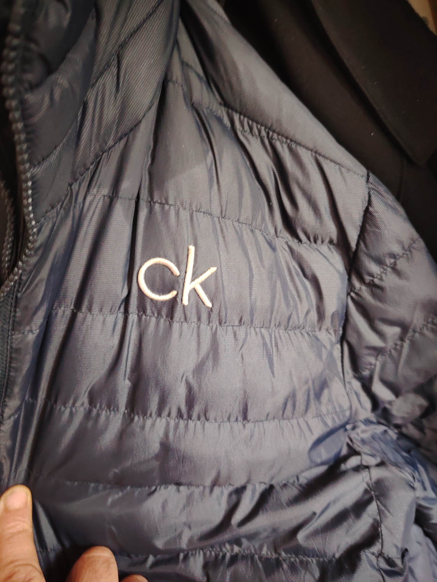 Located in Huntingdonshire, United Kingdom, the image presents a close-up view of a dark blue, horizontally quilted puffer jacket. An embroidered "ck" logo, rendered in light beige thread, is prominently displayed on the upper front of the jacket. A black zipper closure is visible along the left side of the garment. In the lower-left foreground, a human hand, specifically a thumb and parts of two fingers, is lightly grasping the jacket's fabric. The background consists of a dark, indistinct fabric, possibly another garment, situated behind the jacket and to the upper right.