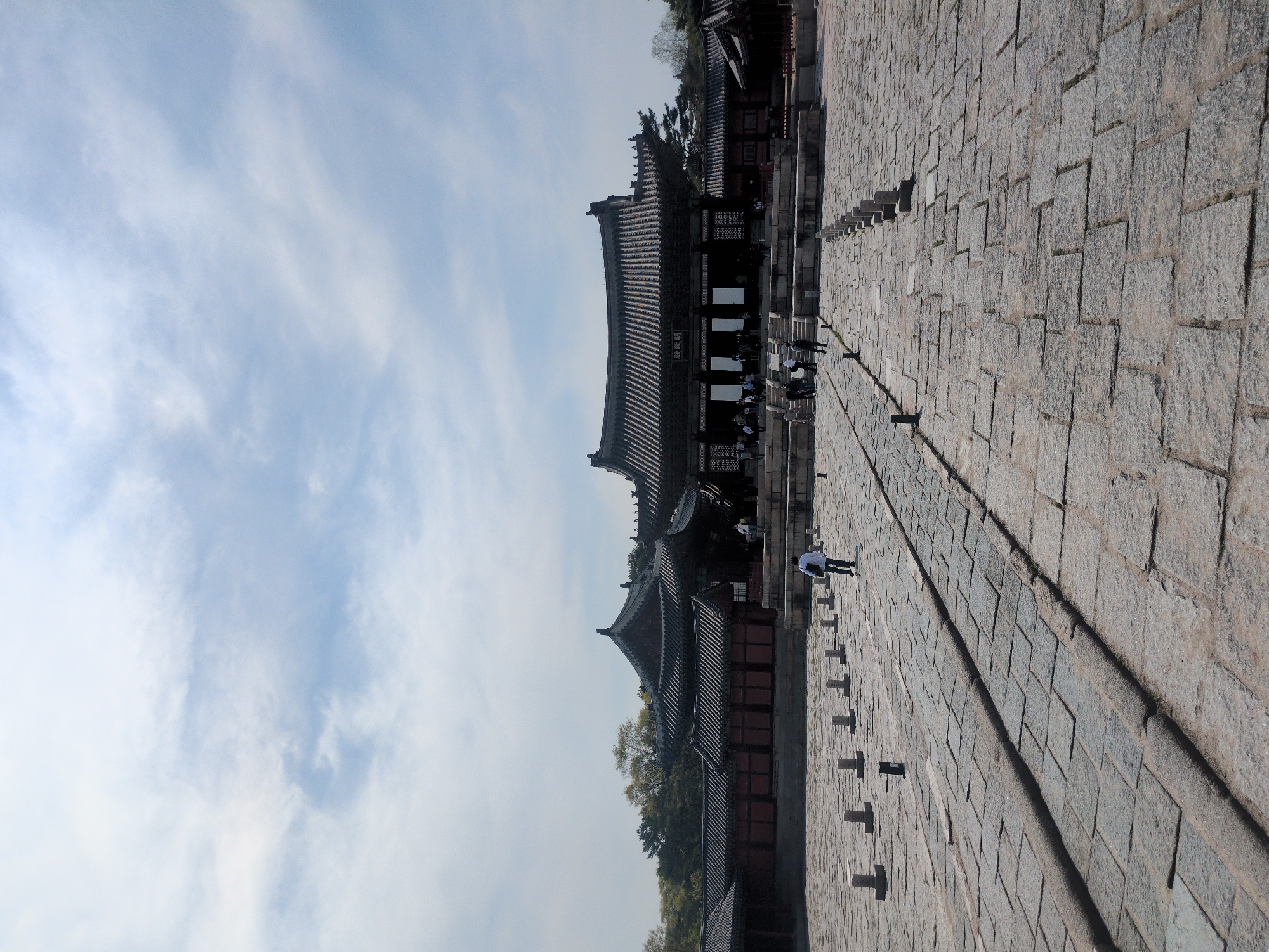 The image shows Changdeokgung Palace, a UNESCO World Heritage Site located in Seoul, South Korea. The building is of traditional Korean architecture. A paved walkway stretches from the foreground, leading towards the palace. Several people are visible near the palace entrance, while one individual is walking along the walkway in the foreground. The sky is visible above the building and is partly cloudy.