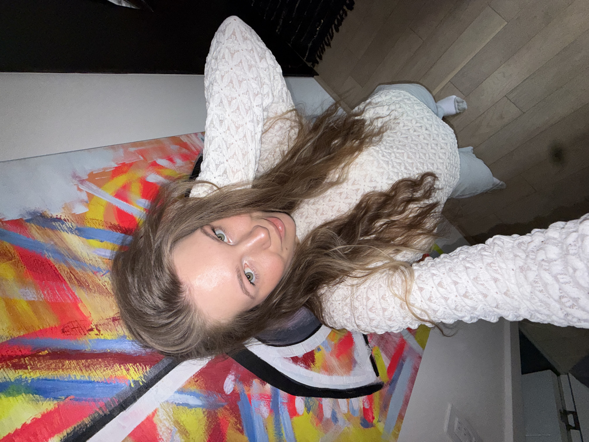 A young woman with light brown hair and green eyes is lying on her side, looking at the camera. She is wearing a white, textured long-sleeved shirt. She is lying on an abstract painting with vibrant colors, including red, yellow, blue, and white. The background includes a wooden floor, a light blue pillow, a white wall, and a black object. The image appears to be taken indoors in a residential setting, possibly in the city of Vozdovac Urban Municipality, Serbia.
