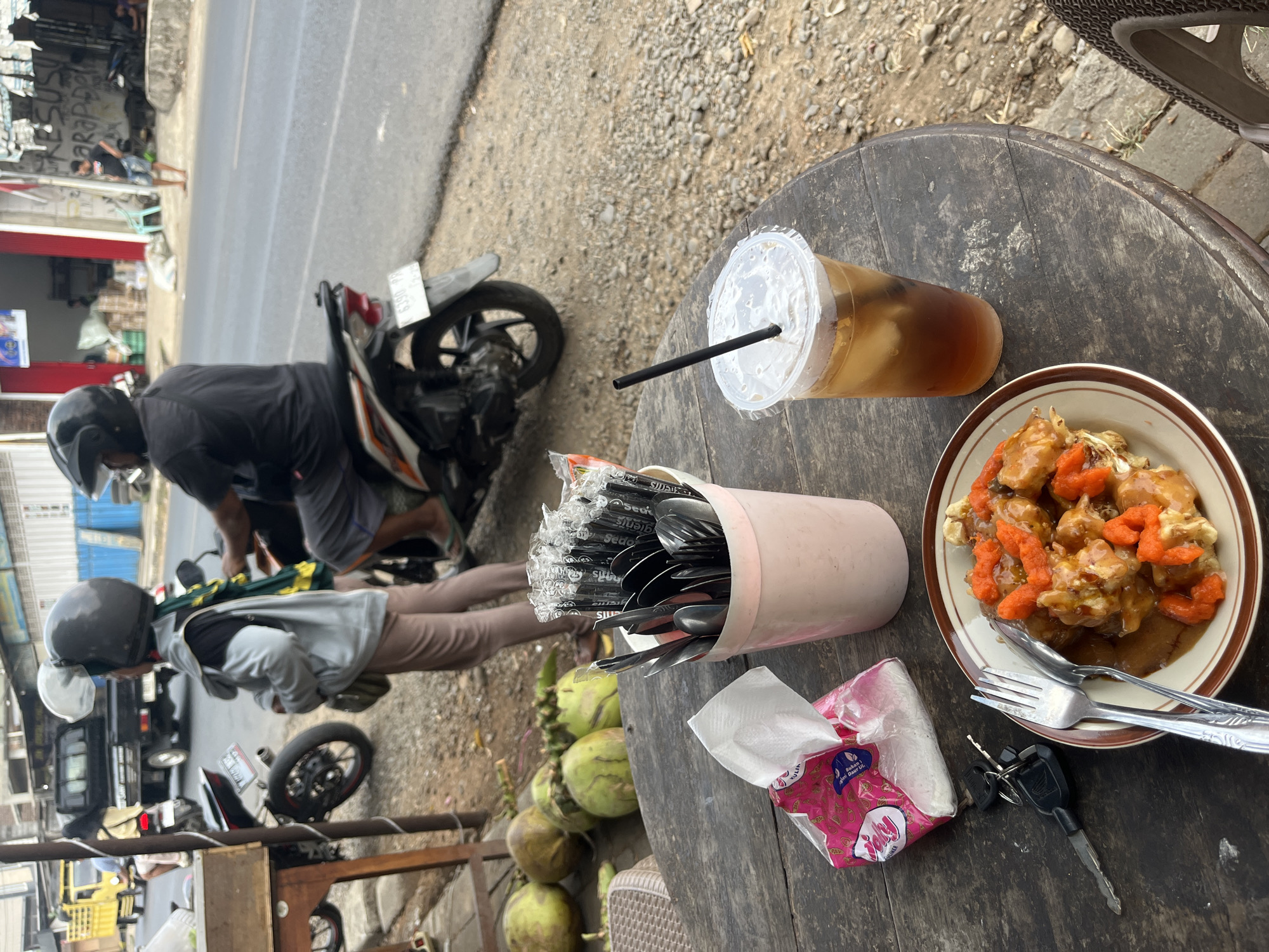 The image captures a scene at a roadside stall in Lebuawu, Indonesia. In the foreground, a wooden table displays a plate of food with orange toppings, a fork and spoon, a packet of tissues, a set of keys, a cup of iced tea with a straw, and a pink container filled with cutlery and straws. In the mid-ground, several coconuts are scattered on the ground. Behind the table, a person in a gray jacket and brown pants stands next to a motorcycle, wearing a helmet. Further in the background, other individuals are visible on motorcycles on a paved road, with buildings lining the street. The environment appears to be a typical urban setting in Indonesia, with motorcycles and local commerce being prominent.
