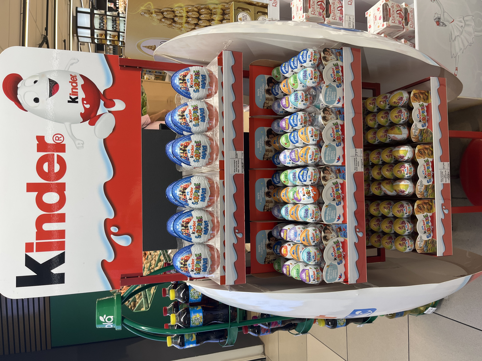 The image shows a large, multi-tiered display of Kinder Surprise eggs in a retail environment, likely a supermarket. The display is shaped like a large egg and features the Kinder logo prominently at the top, along with an illustration of the Kinder egg mascot.
The display has several shelves filled with different varieties of Kinder Surprise eggs, individually wrapped in colorful foil. The eggs are arranged in rows and columns, showcasing various designs and packaging. Some of the eggs have images of characters from popular franchises like Disney, as indicated by the text and graphics on their wrappers.
Below the Kinder display, there are also boxes of Raffaello chocolates stacked on a shelf to the right. In the background, shelves with other products are visible, as well as part of a beverage display with bottles of soda. The flooring appears to be tiled, typical of a retail setting.
The lighting suggests an indoor environment with artificial illumination. No people are clearly visible in the image, though a blurred figure can be seen in the background. The overall scene depicts a commercial setting focused on the sale of confectionery products.
