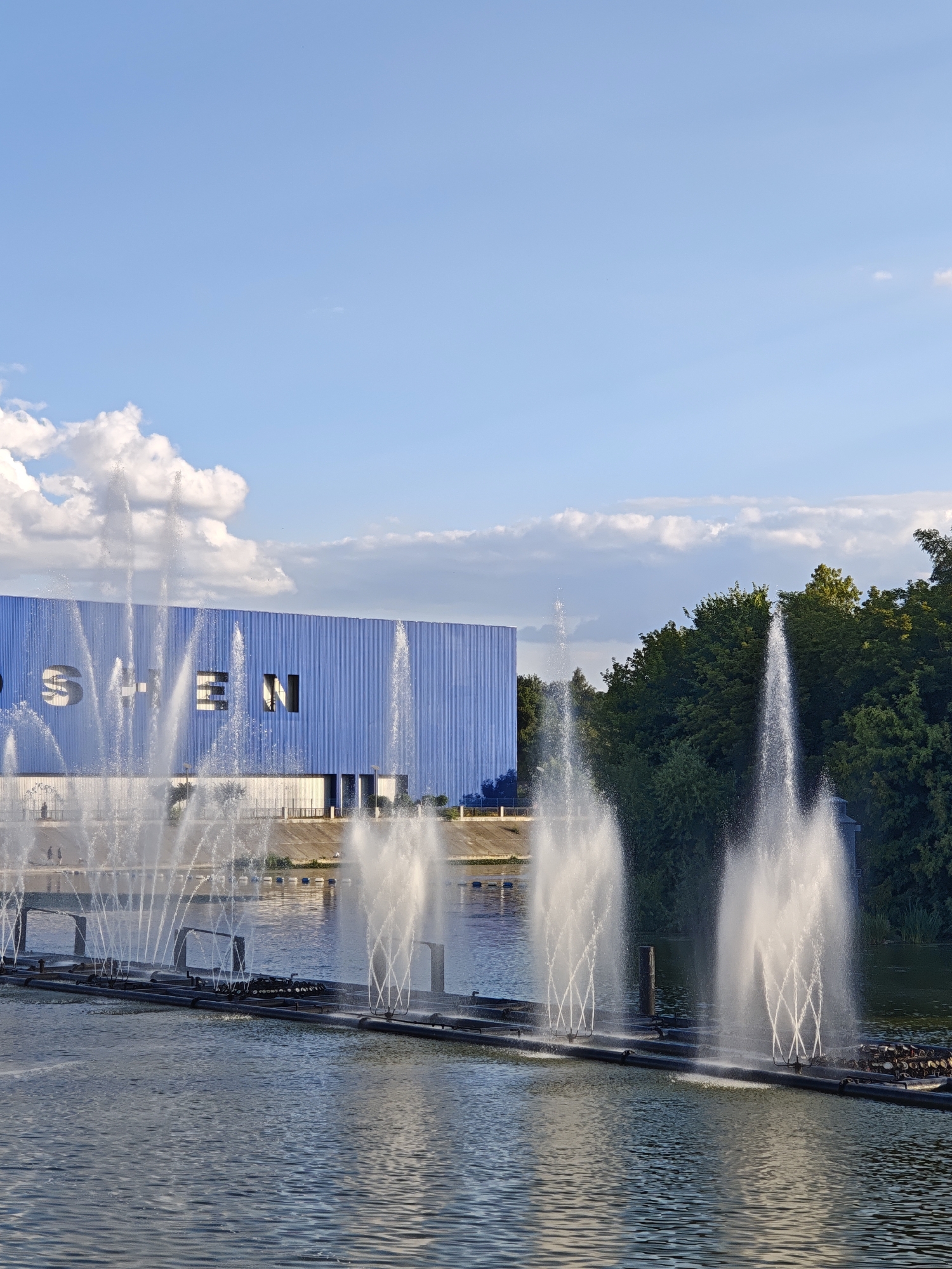 The image depicts a scenic view of the Roshen Fountain in Vinnytsia, Ukraine. In the foreground, a large body of water is disturbed by numerous water jets from a fountain, creating a dynamic visual effect. The jets vary in height and pattern, with some forming delicate cascades and others erupting in powerful plumes. In the background, a large, modern building with a blue facade stands prominently. The building features large, stylized letters that appear to spell "ROSHEN". The sky above is a clear blue, dotted with fluffy white clouds, indicating fair weather. Lush green trees line the far bank of the water, providing a natural contrast to the urban architecture. The overall scene captures a lively and impressive urban landscape, highlighting the impressive engineering and visual artistry of the fountain.