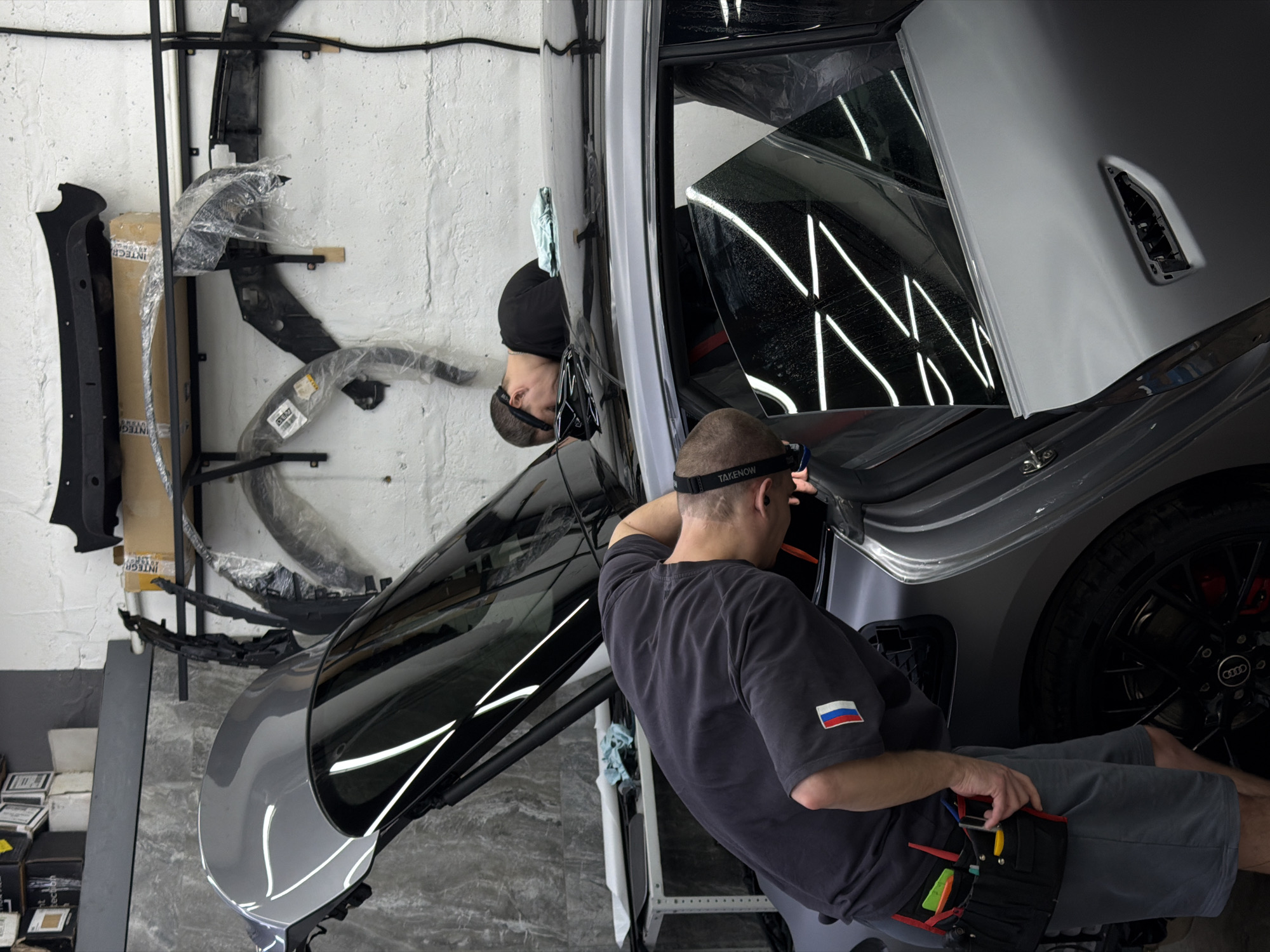 Two men are working on a gray car in a garage. One man is sitting inside the car, facing the front, and wearing a gray t-shirt with a Russian flag patch on the sleeve. He has a black tool belt on his lap. The other man is standing outside the car, leaning in, and wearing a black cap. The car has its door open, and the inside of the door is covered in a patterned film. Various car parts are hanging on a metal rack against a white wall in the background. Boxes are stacked on the floor to the left of the rack. The location appears to be Krasnoyarsk, Russia, indicated by the Russian flag patch on the man's shirt.