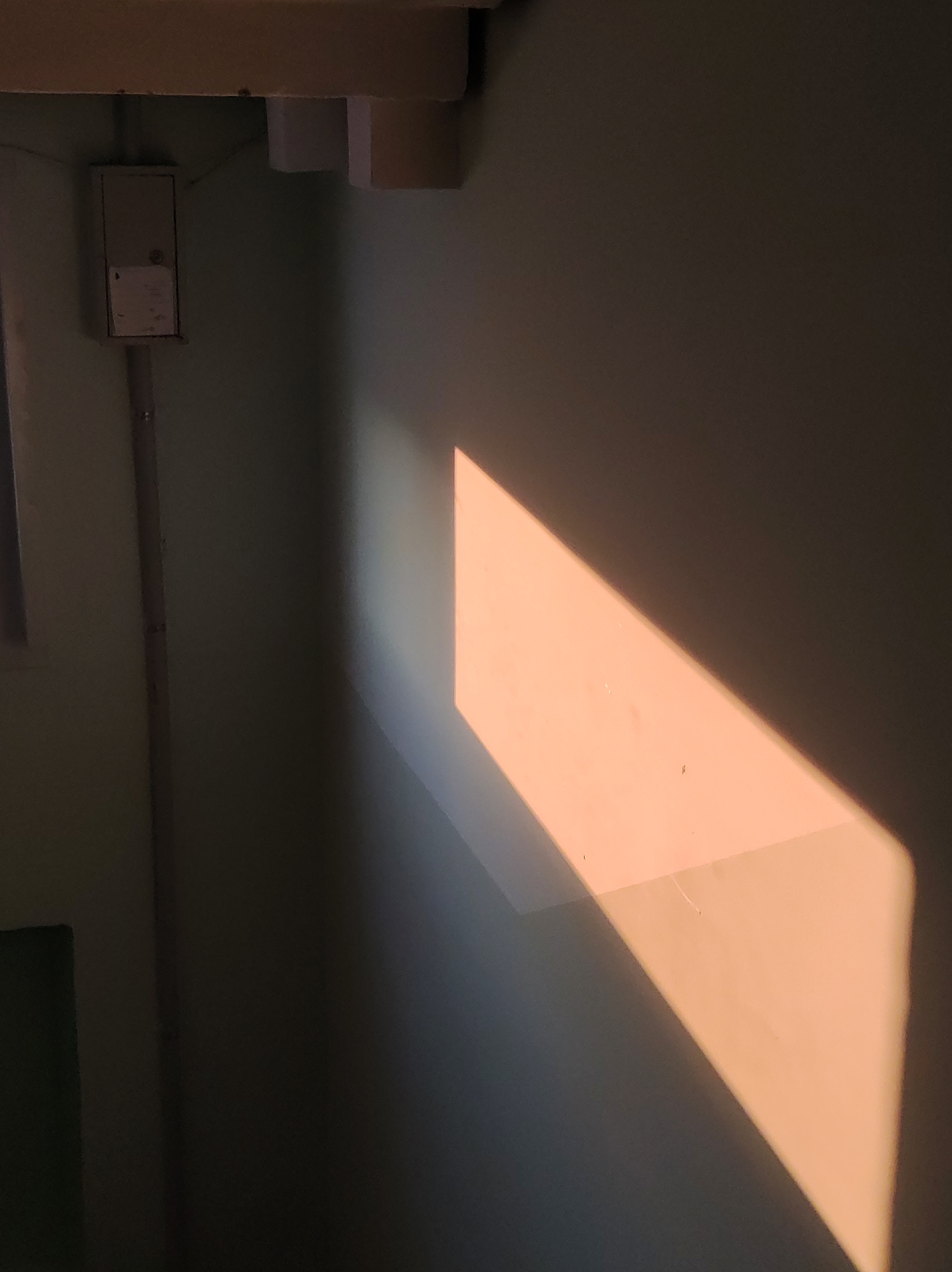 The image presents an interior corner, likely a room, with light casting a diagonal, trapezoidal shape on the wall. To the left of the frame, electrical conduits and a junction box are affixed to the wall. The light source is not visible, but its illumination creates a contrast between light and shadow. The wall appears to be painted a muted color. There are no discernible people or specific identifiable objects in the image to place it in a city or location.