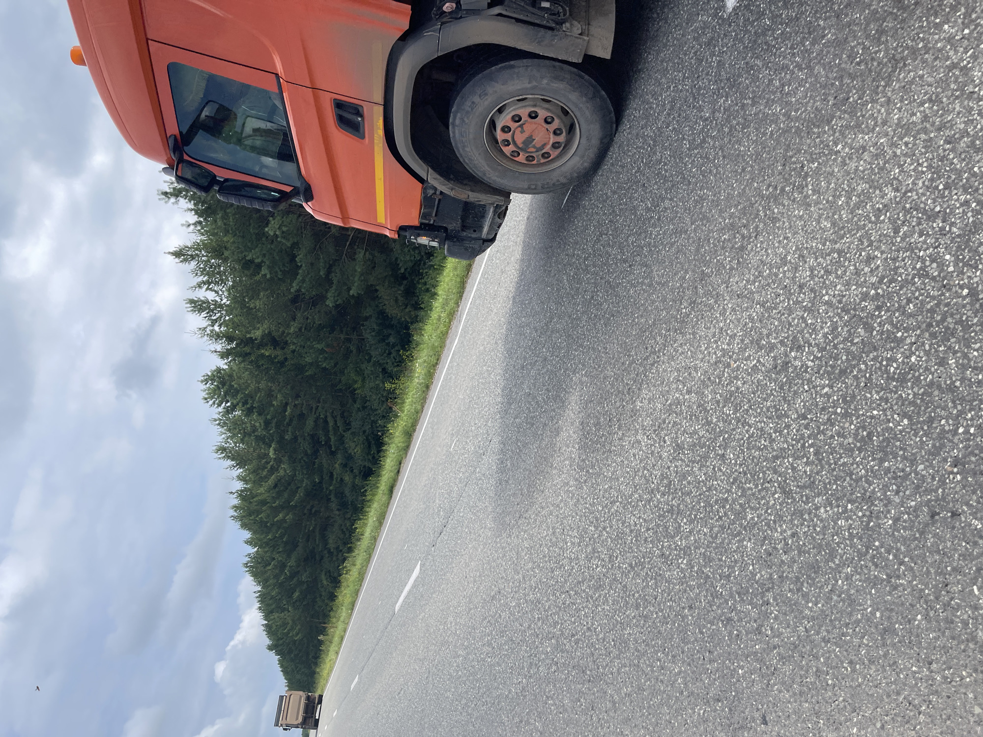 An orange truck is parked on the right side of a two-lane asphalt road, partially obscuring the foreground. The truck is facing towards the left of the frame, and only its front passenger side is visible. A second, smaller brown truck is visible in the distance on the left side of the road, also traveling towards the left. The road is bordered by a dense forest of dark green pine trees on the right and left. The sky is overcast with scattered white clouds. The scene appears to be located in Perevoznoe, Russia, on a rural road.