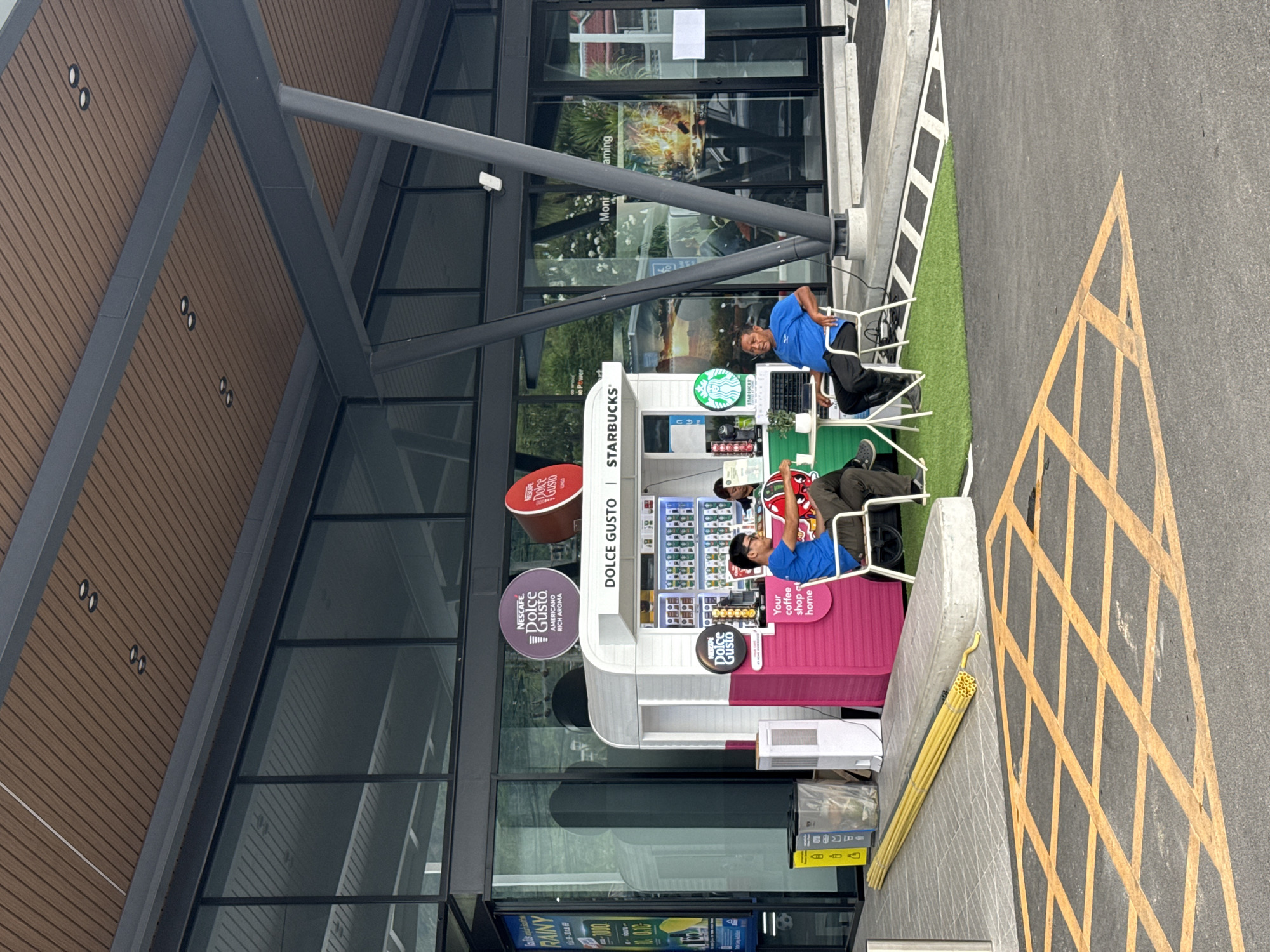 The image depicts an outdoor Starbucks coffee kiosk located in Baan Chaweng Noi, Thailand. Two men are seated at a small table on a patch of artificial grass in front of the kiosk. The kiosk itself is a bright pink and white structure with a variety of coffee products displayed inside. Behind the kiosk, the facade of a building with large glass windows is visible, reflecting the surrounding environment. The ground in the foreground is paved asphalt with yellow road markings. The overall scene suggests a commercial area with a focus on casual outdoor seating and beverage service.