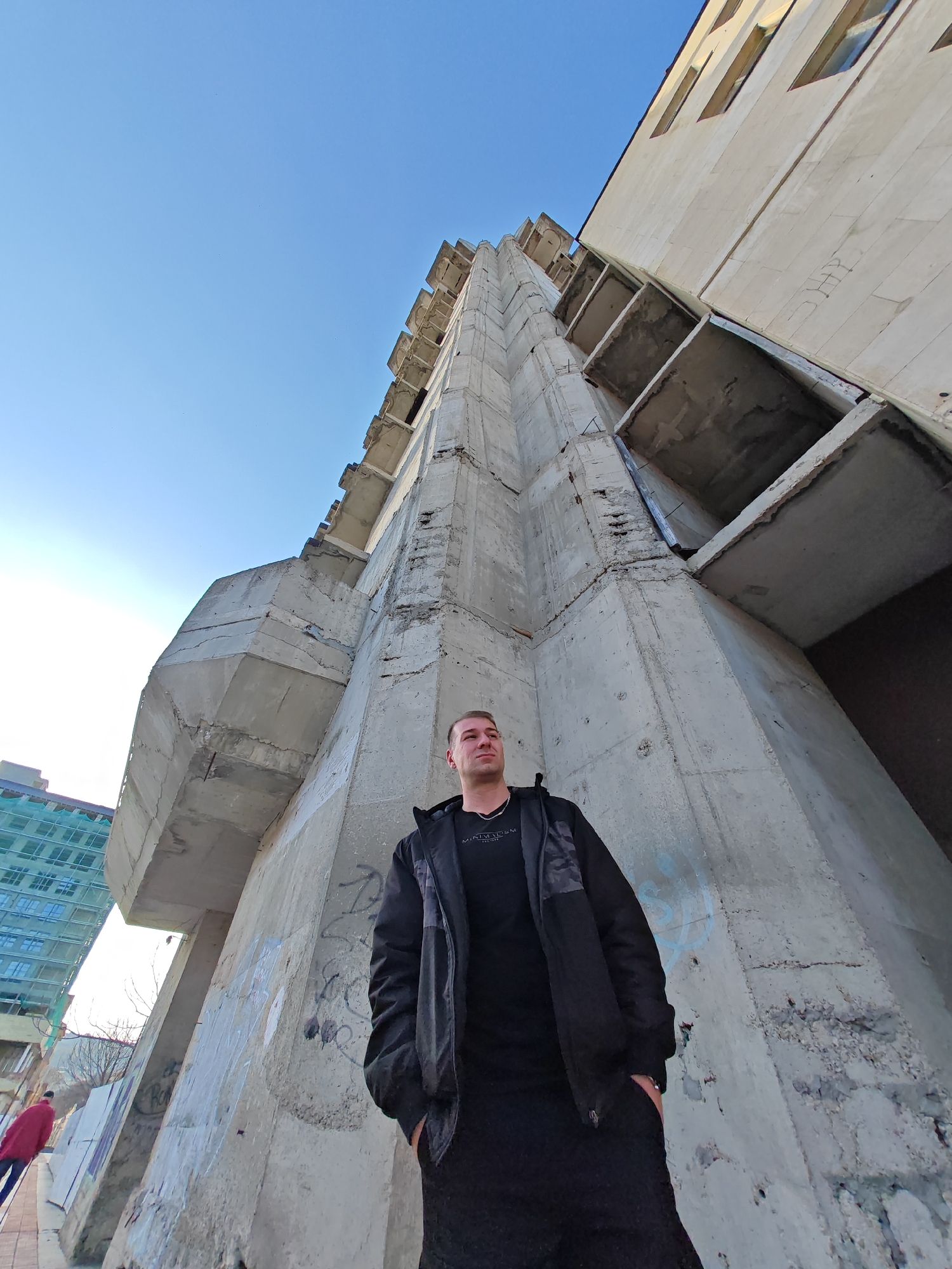 An adult male stands in the lower center of the frame, looking upwards and to his right, positioned in front of a prominent, multi-story, raw concrete brutalist structure identified as the unfinished House of Culture building in Shumen, Bulgaria. The male, Caucasian with short brown hair, wears a black t-shirt with white text, a black padded jacket with grey shoulder accents, and black trousers, with hands in pockets. The concrete structure exhibits exposed rebar, surface weathering, cracks, and graffiti, including blue spray paint near its base. It features numerous unglazed, rectangular openings resembling balconies or windows. To the right and behind this, a taller, beige concrete building with recessed rectangular windows extends upwards. On the far left, a modern building with a blue glass facade is partially visible, appearing to be under construction or renovation. A distant, indistinct figure clad in red walks on a paved sidewalk on the far left. The scene is illuminated by bright daylight under a clear blue sky.