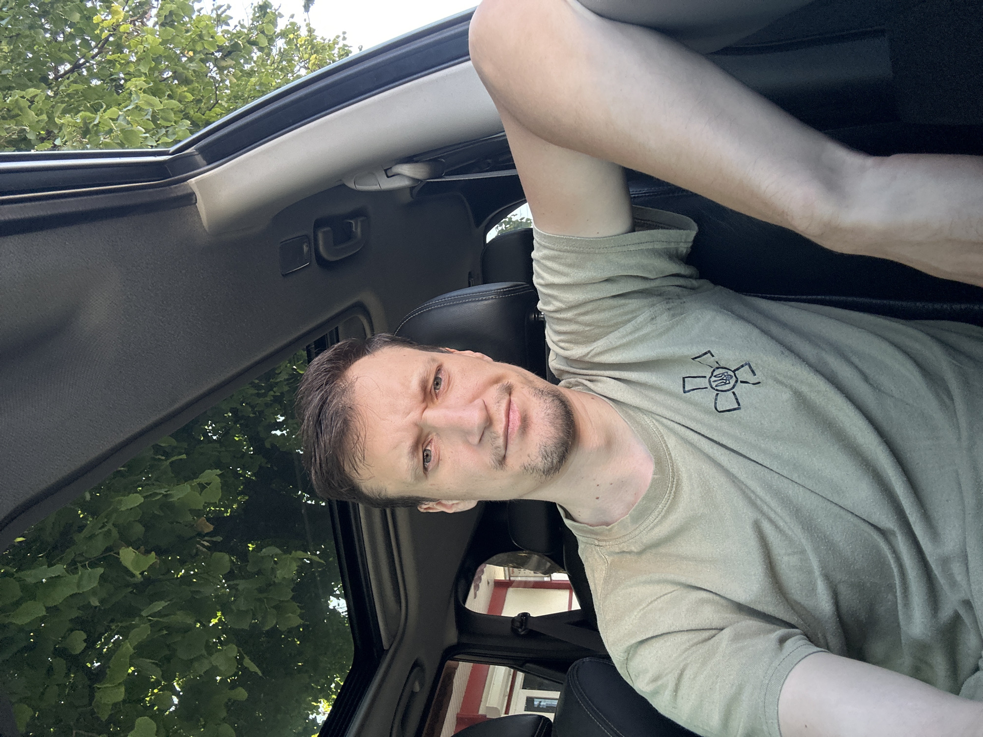 A man is pictured inside a car, seated in the driver's seat. The car's sunroof is open, allowing a view of green trees and foliage above. The man is wearing a light brown t-shirt with a black emblem on the left chest. His left arm rests on the center console of the car. The interior of the car appears to be black leather. The background outside the car window shows the facade of a building with red trim and windows, possibly in a city setting like Kamianets-Podilskyi, Ukraine. The lighting suggests it is daytime, with sunlight filtering through the trees.
