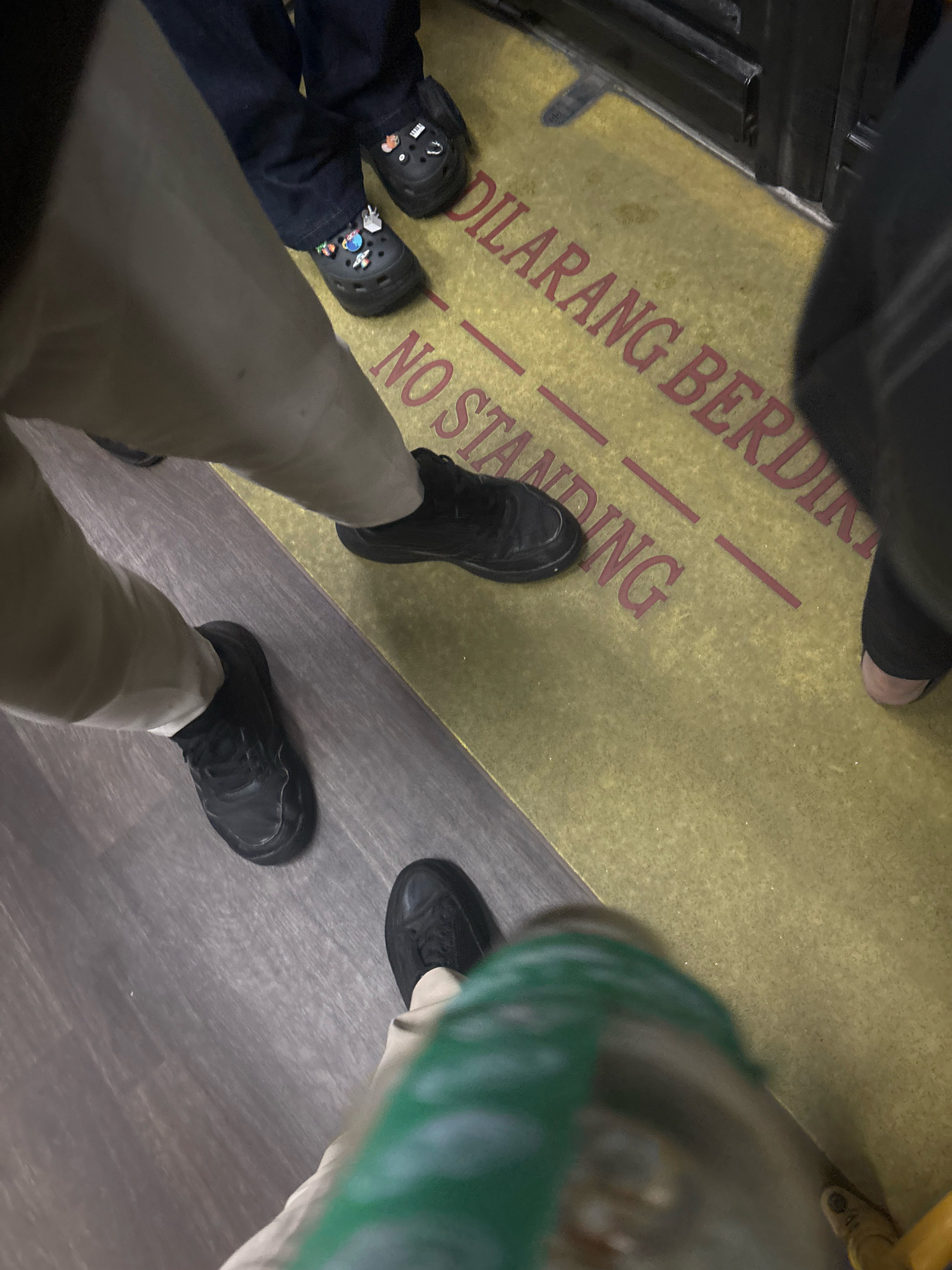 The image shows the lower bodies of several people standing in what appears to be a public transport vehicle, likely a bus. The floor is covered with a yellow mat that has red lettering. The text on the mat reads "DILARANG BERDIRI" and "NO STANDING", indicating a rule against standing. One person is wearing black Crocs with various decorative charms. Other individuals are wearing black sneakers. The flooring of the vehicle is divided between the yellow mat and a grey, wood-patterned surface. The scene is shot from a low angle, focusing on the feet and legs of the individuals. Based on the text in Indonesian, "DILARANG BERDIRI," which translates to "Standing Forbidden," and the presence of other people in what appears to be a public transportation setting, this scene is likely within a public transport vehicle in North Jakarta, Indonesia, adhering to local regulations.