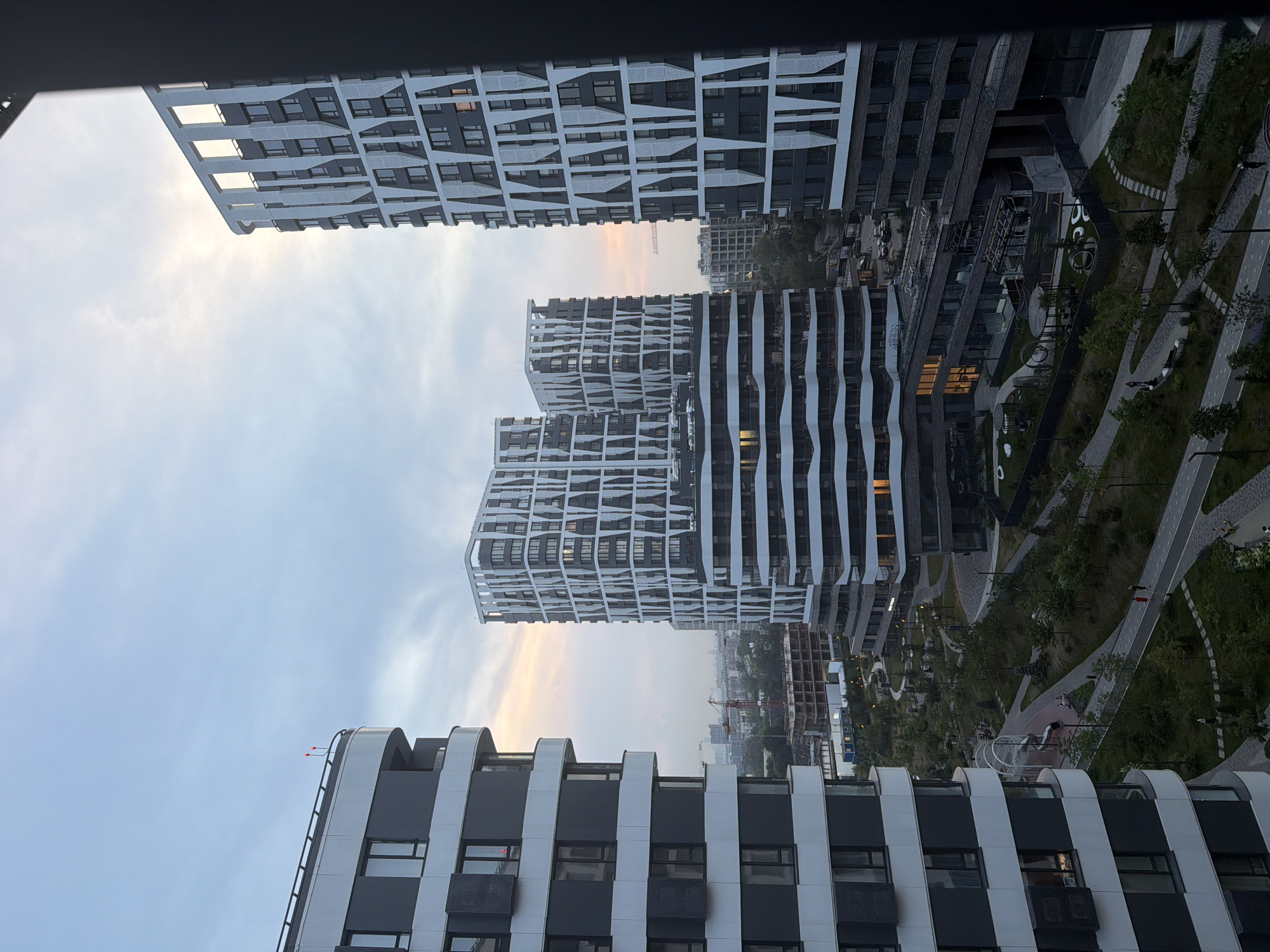 This image captures a contemporary urban landscape in Moscow, Russia, likely within a newly developed residential complex. The scene is dominated by several modern apartment buildings with striking architectural designs. One building on the left features a curved facade with alternating dark grey and white horizontal bands, punctuated by dark windows. To the right, a cluster of taller buildings exhibits a more geometric aesthetic, with white, angular patterns overlaying dark facades. These buildings feature numerous windows, some illuminated, suggesting occupancy.
Below the residential towers, a well-maintained park area is visible, complete with winding pathways, landscaped greenery, trees, and outdoor recreational spaces like a playground with a slide. A few individuals are present in the park, walking along the paths or sitting on benches, indicating a functional and inhabited community space. In the background, other urban structures and construction sites, including a crane, are discernible, signifying ongoing development in the city. The sky above is a soft gradient of blue and pale orange, hinting at either dawn or dusk, and casting a diffused light over the scene. The overall impression is one of modern urban living with integrated green spaces.