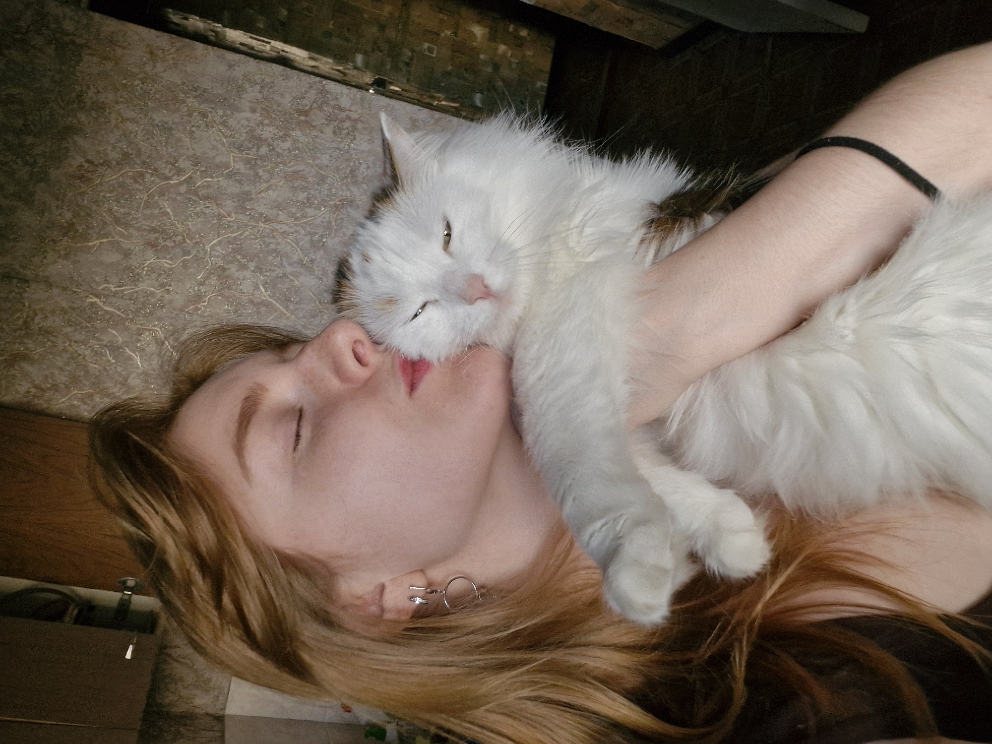 A woman lies on a surface with a textured pattern, possibly a bed or the floor, with a long-haired white cat cradled in her arms and its head resting on her face. The woman has red hair and closed eyes, and is wearing earrings. The cat has eyes partially open. The location is not discernible from the image and it is impossible to determine if it's in Yekaterinburg, Russia.
