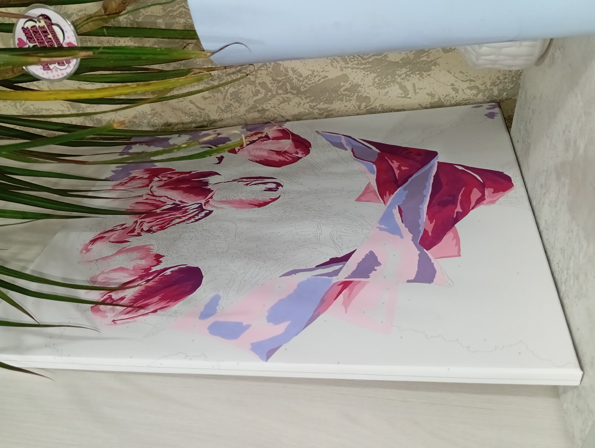 The image shows a partially painted paint-by-numbers canvas depicting red and purple flowers, possibly tulips. The canvas is white with numbered sections. Some sections are painted in various shades of red and purple. The canvas is lying flat on a surface that appears to be a table or shelf, with the corner of the table visible in the lower right. Green, leafy plant stems lie across the left side of the canvas. A round, white label with some text and a pink design is visible amidst the plant stems. The background is a beige and white patterned wall. Above the canvas, there is a white or light-colored object that is not fully visible, but might be part of the ceiling or another object leaning against the wall. The photograph was taken in the city of Yurgamysh, Russia.