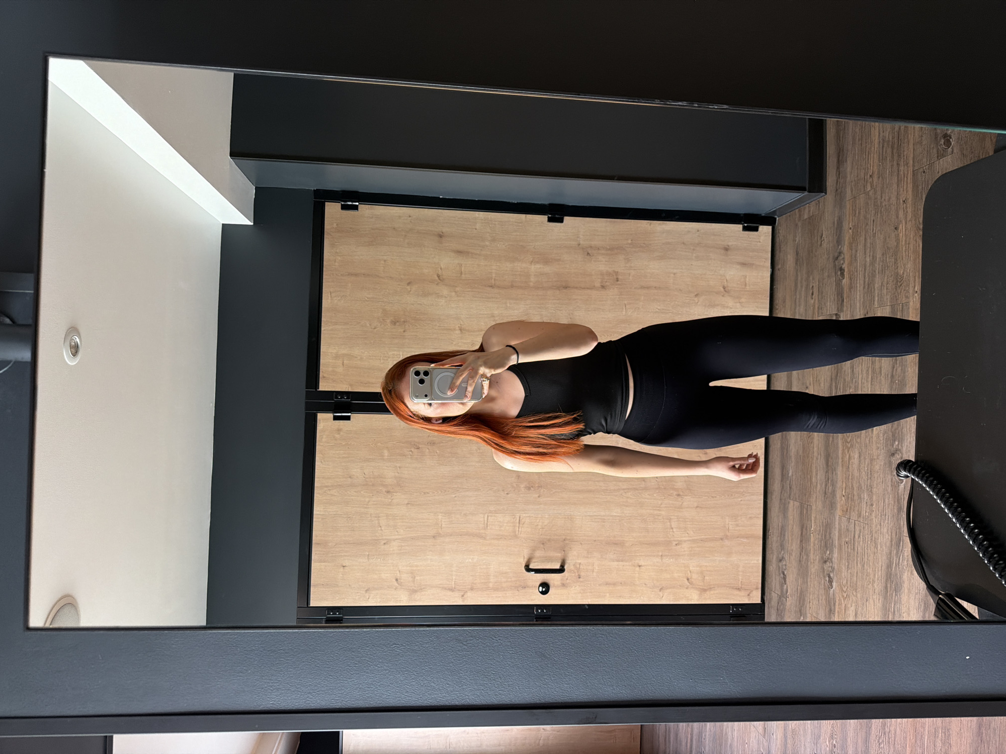A woman with red hair is taking a selfie in a full-length mirror. She is wearing a black tank top and black leggings. The background includes a beige door with black hardware, a light-colored wall, and a black wall. There is a black chair visible on the right side of the mirror's reflection. The location of the image is not possible to determine based on the available visual information.