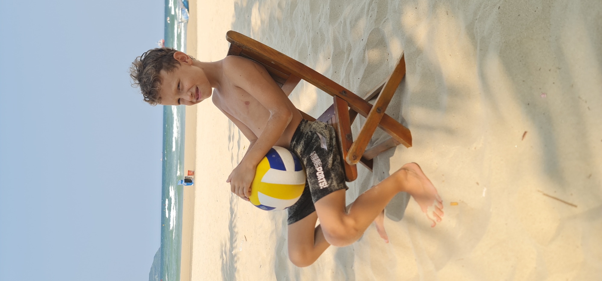 This image depicts a playful moment on a sunny beach in Đà Nẵng, Vietnam. The main subject is a young boy, appearing to be in elementary school, who is shirtless and wearing dark swim shorts with the white text "TOUGH SPORTS!" visible on them. He has light brown, curly hair and a subtle smile or smirk on his face, suggesting contentment or amusement.
The boy is positioned kneeling or crouching on the light-colored sand, leaning against a rustic wooden folding beach chair that is also on the sand. He holds a yellow, blue, and white volleyball in his arms. The background features a vast expanse of the ocean with gentle waves breaking on the shore, beneath a clear, light blue sky. The bright conditions indicate it is daytime, likely mid-morning or afternoon, with shadows cast on the sand by the chair and possibly overhead foliage, enhancing the sense of a warm, bright day. The overall scene conveys a relaxed and enjoyable beach atmosphere.